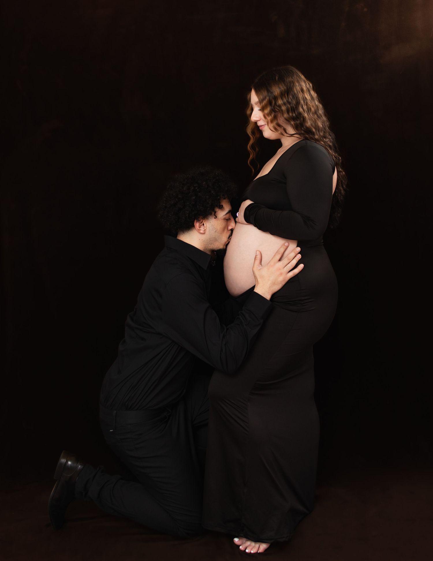 Dramatic maternity photos of couple in black attire against dark backdrop.