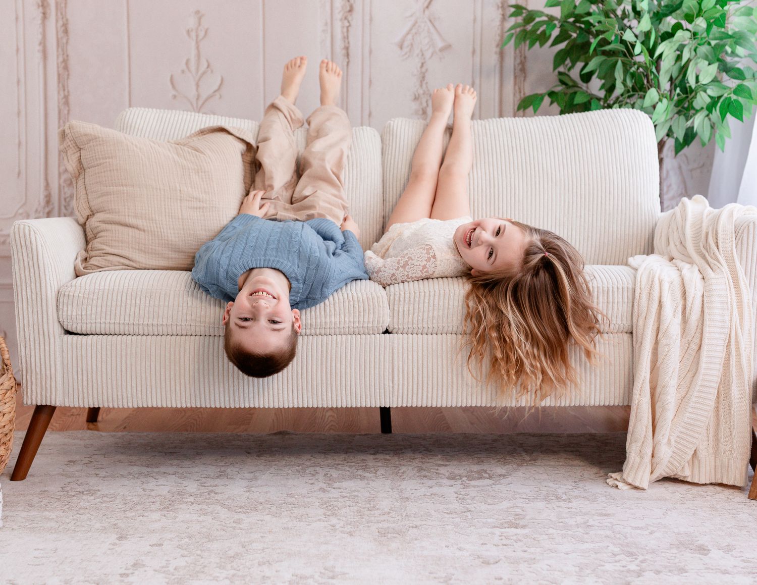 Two people playfully lounging upside down on a beige couch with a throw blanket and plant in the background.