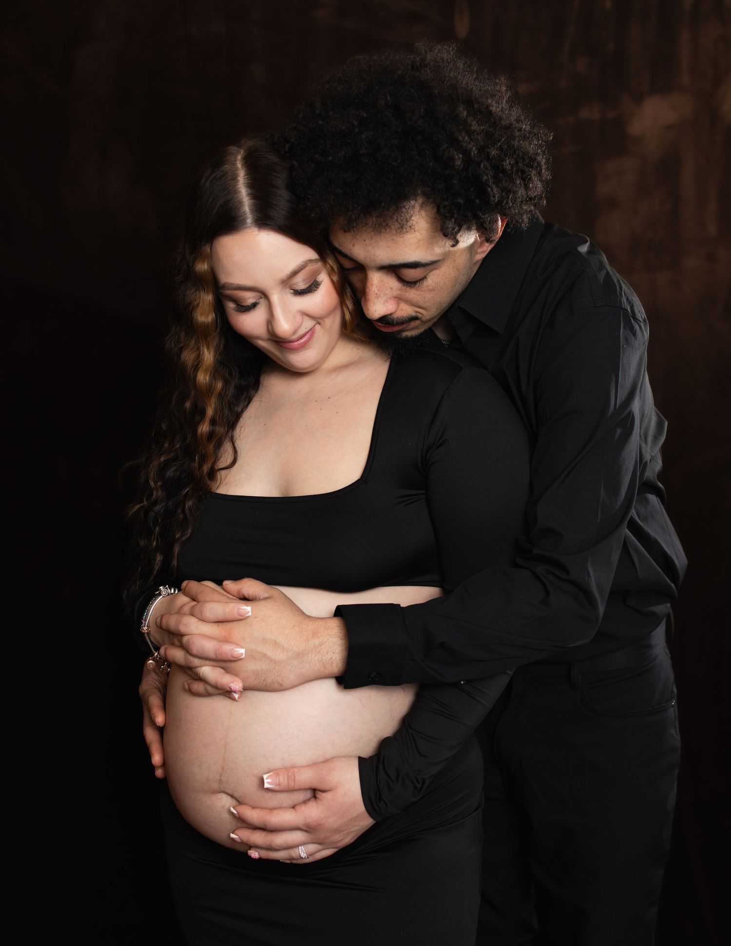 A couple in black clothing shares an intimate maternity photoshoot moment against a dark background.