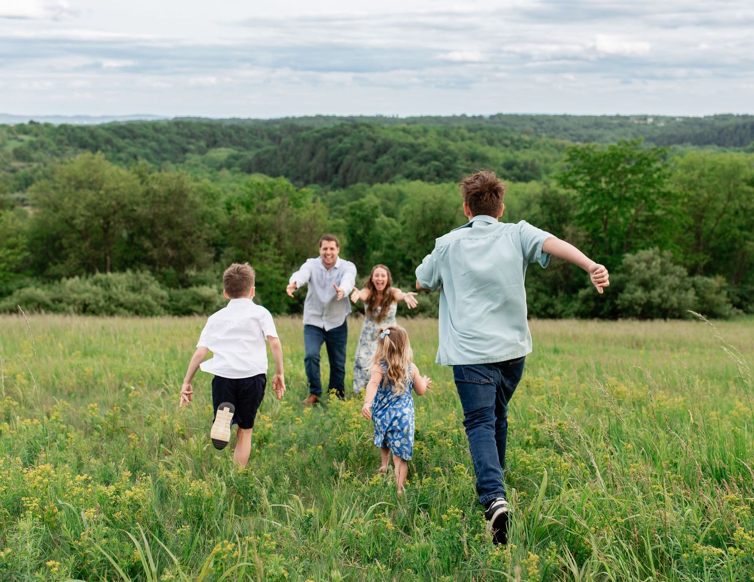 Family running playfully through wildflower meadow with hills in background.