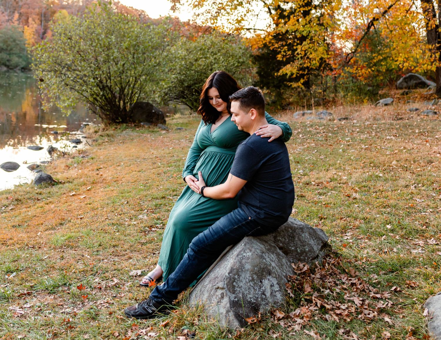 Couple poses on large rock during fall maternity photoshoot near water.