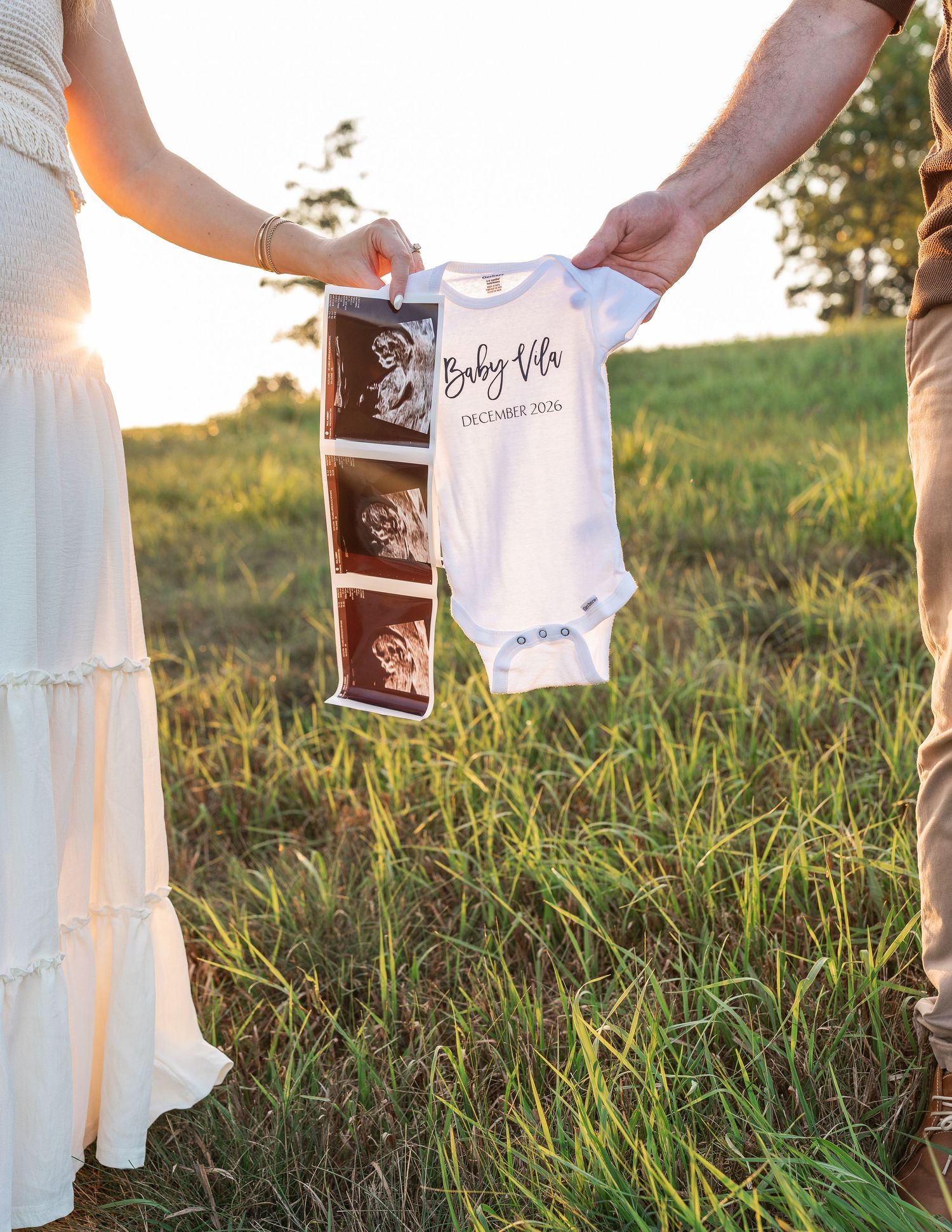 Couple holding ultrasound photos and a Baby Vila onesie in a sunny green field for a pregnancy announcement.