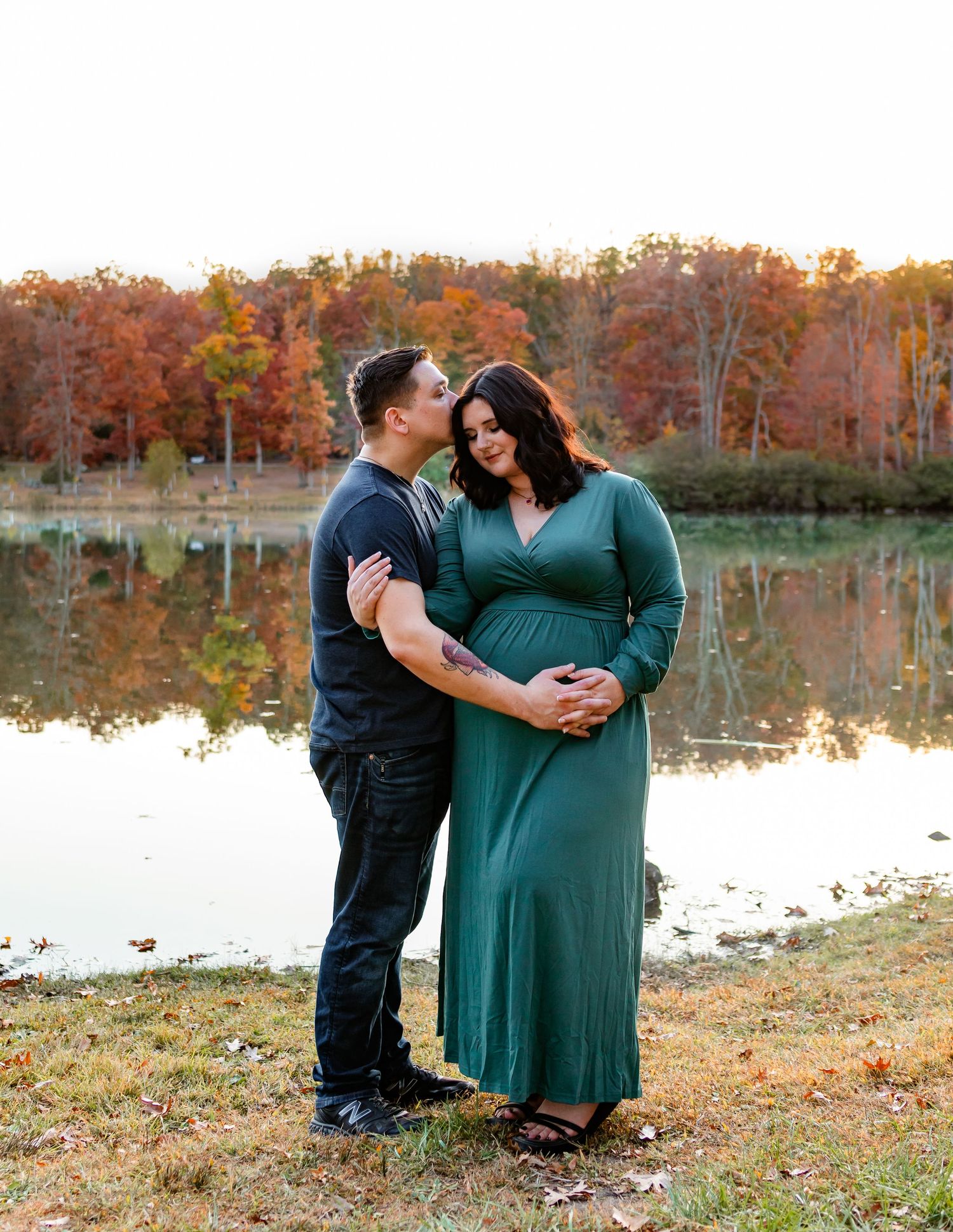 Autumn maternity photoshoot by lake with couple in green dress and black attire.