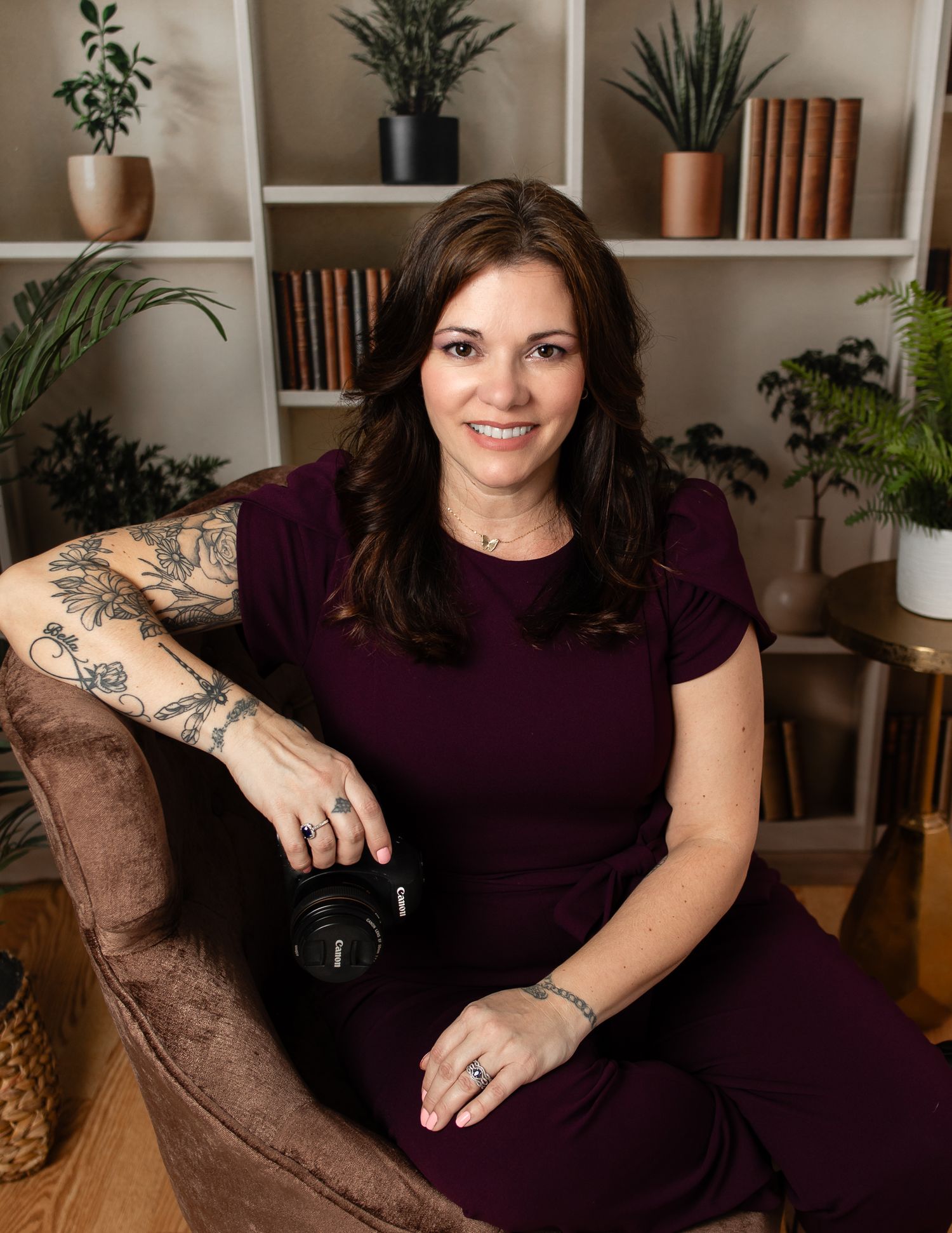 Woman in purple top sits on brown couch with indoor plants and bookshelves in background.