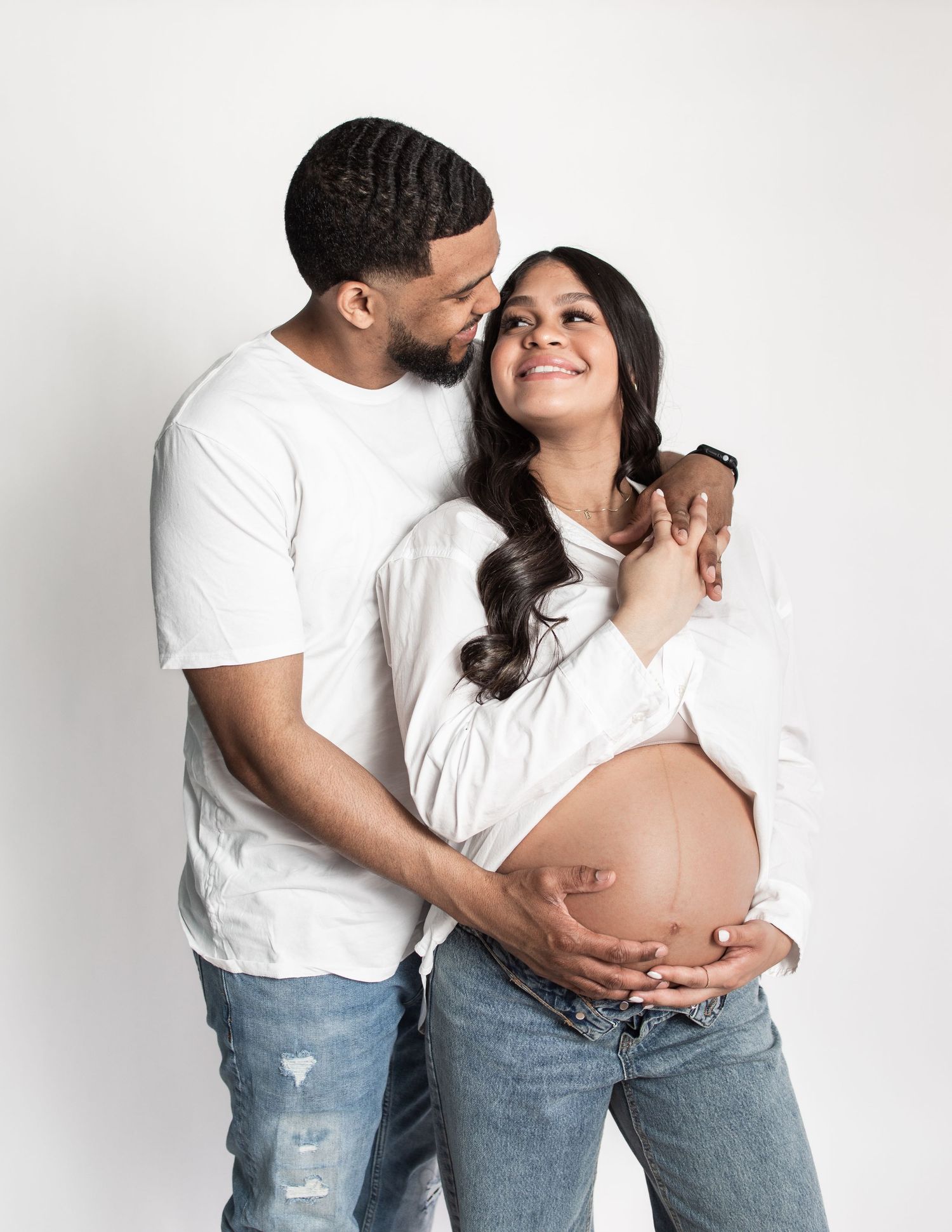 Expectant couple in casual white tops and jeans pose together against a white background.