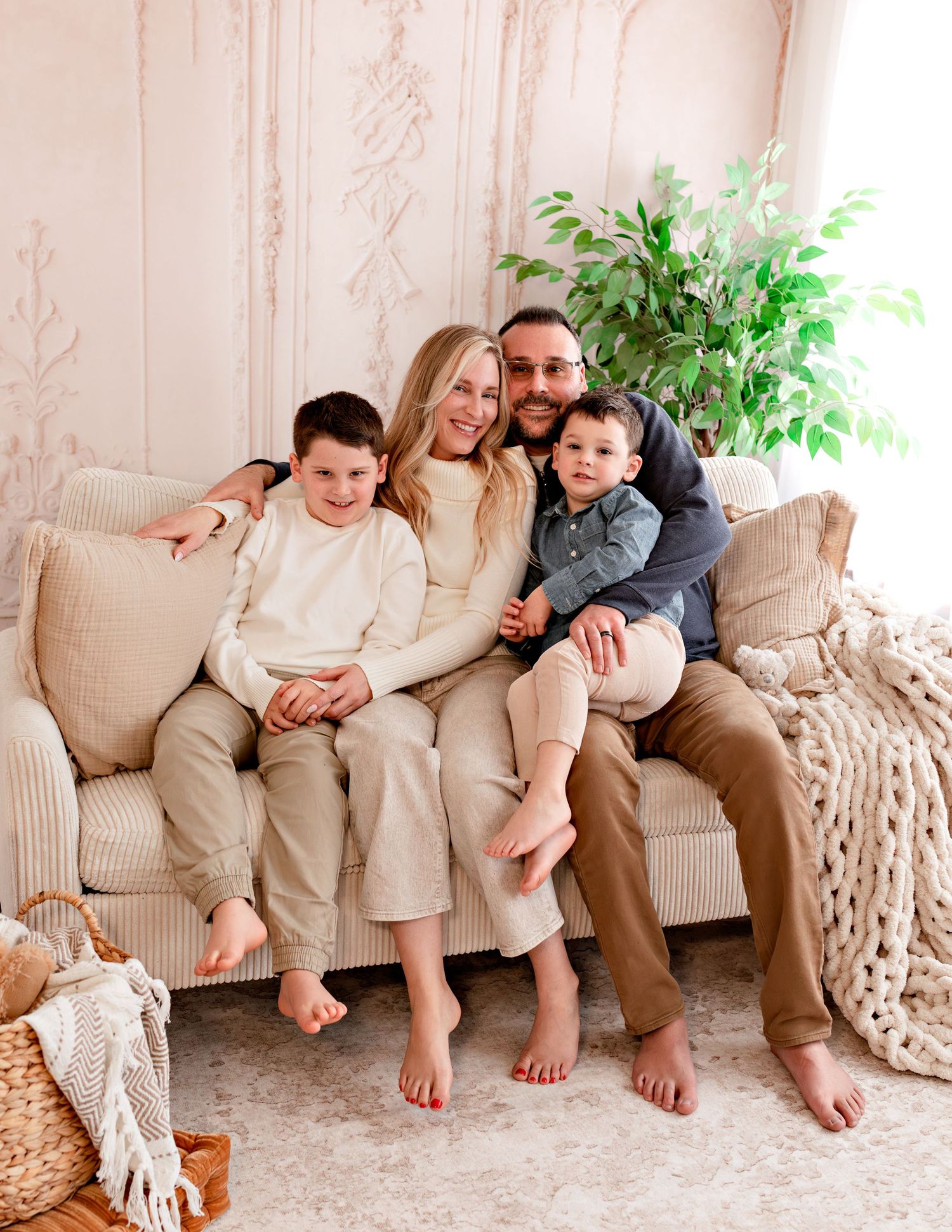 A happy family of four sits barefoot on a beige couch in a cozy, neutral-toned living room.