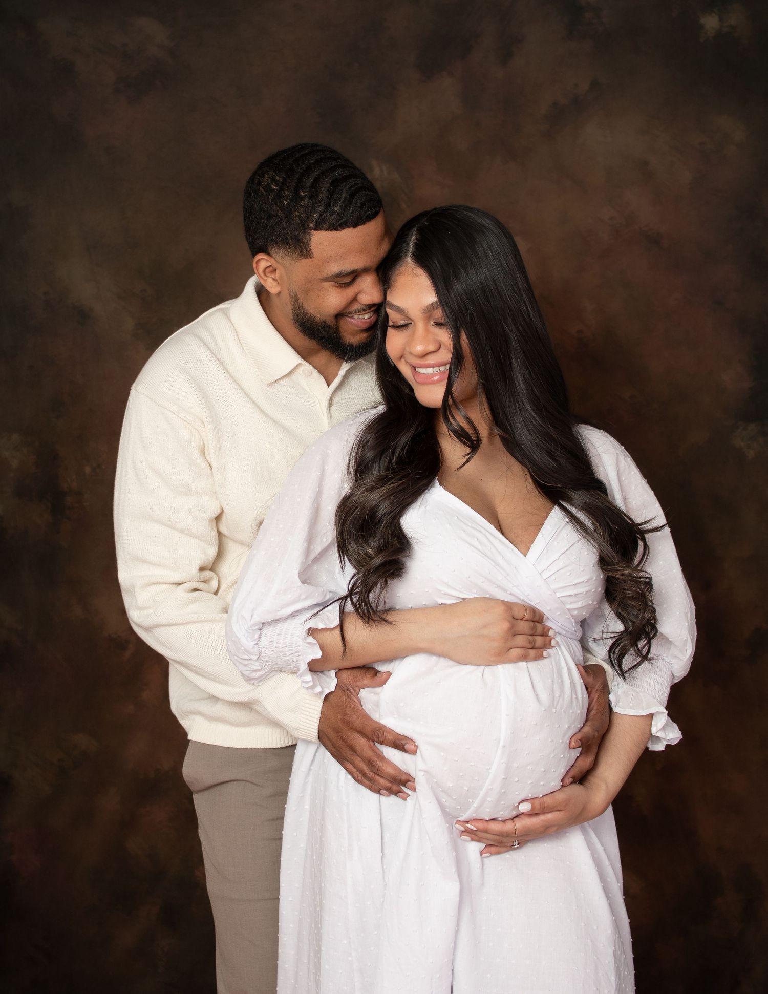 Romantic maternity photoshoot with couple in white outfits against moody brown background.