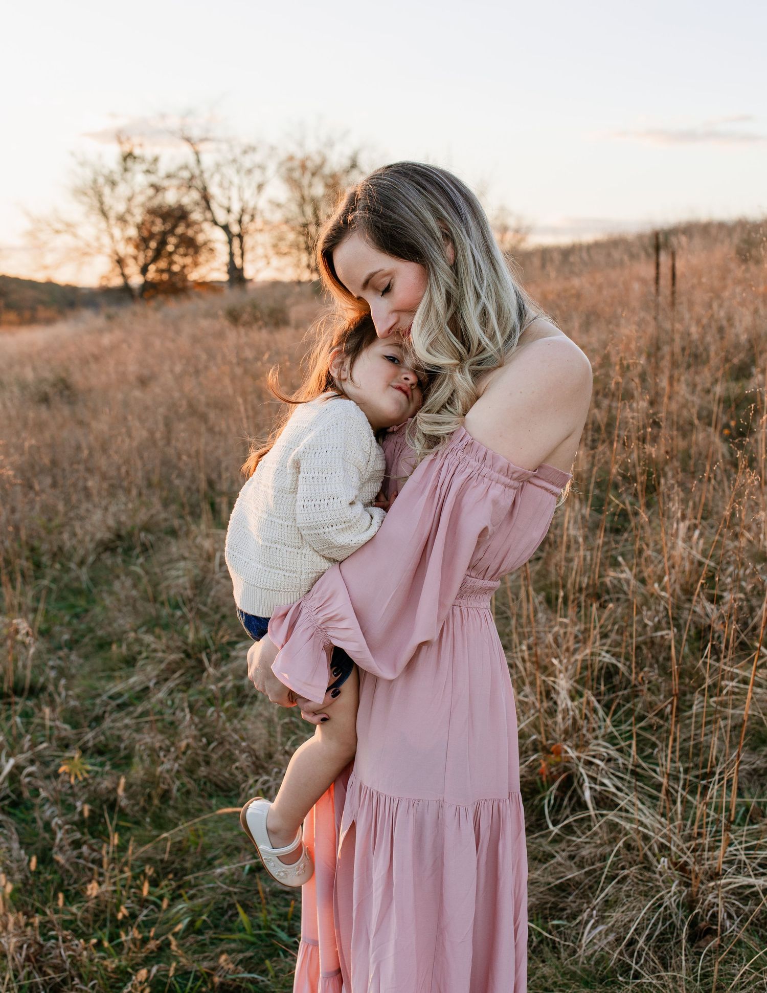 Mother and child share tender moments in a sunlit field at golden hour wearing pink and white dresses.