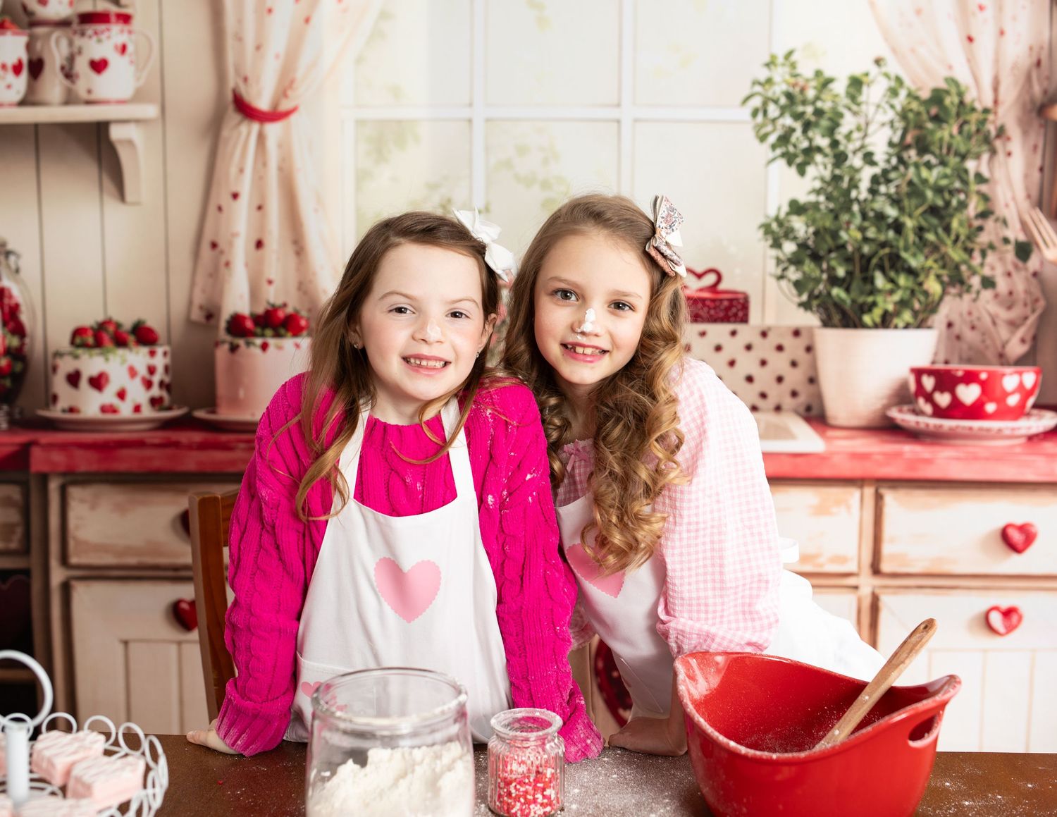 Two children in white aprons stand together in a Valentine's Day themed kitchen setting with red and pink decor.
