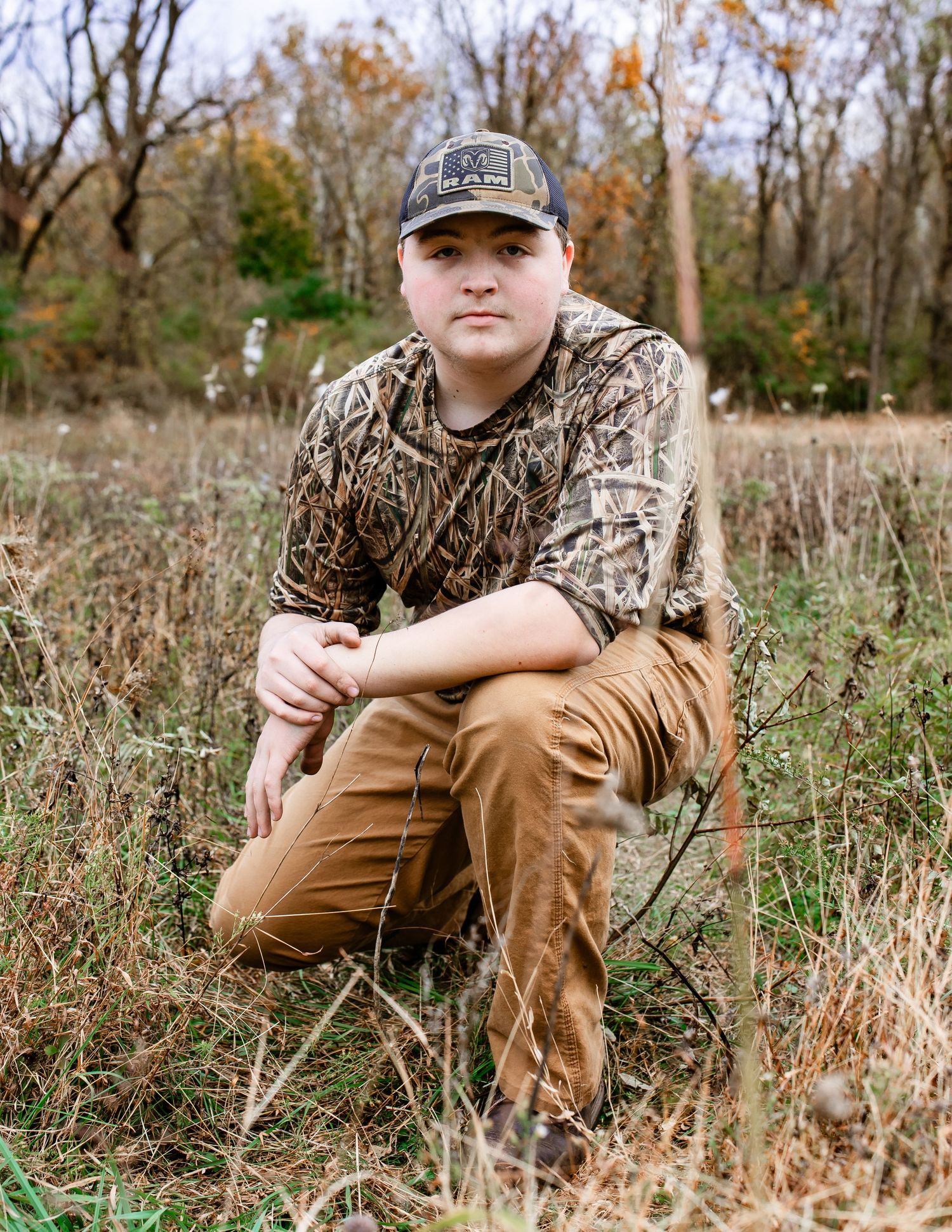 Young hunter in camouflage clothing and brown pants sits in autumn field.