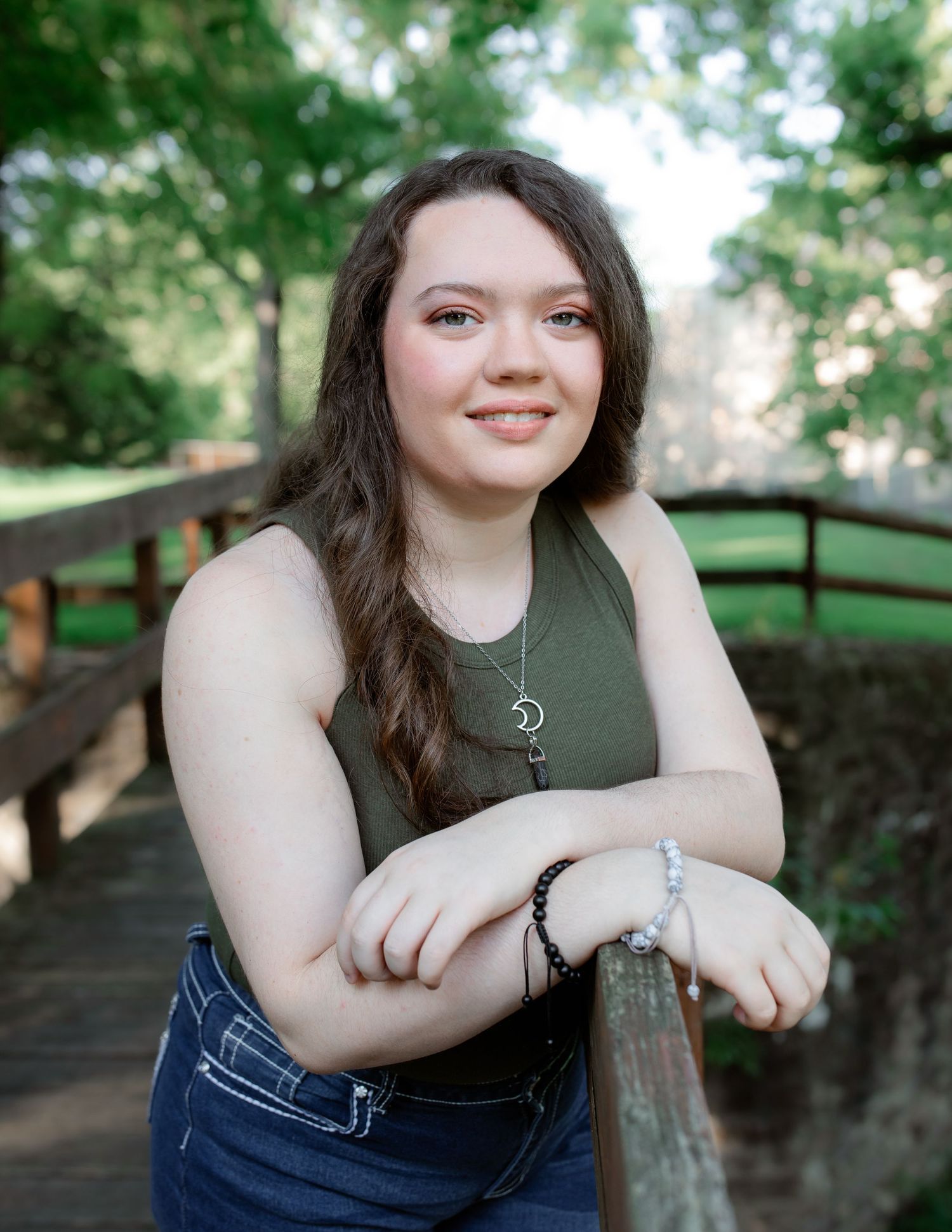 A person in a green tank top and jeans poses on a wooden bridge in a lush outdoor setting. Senior Session