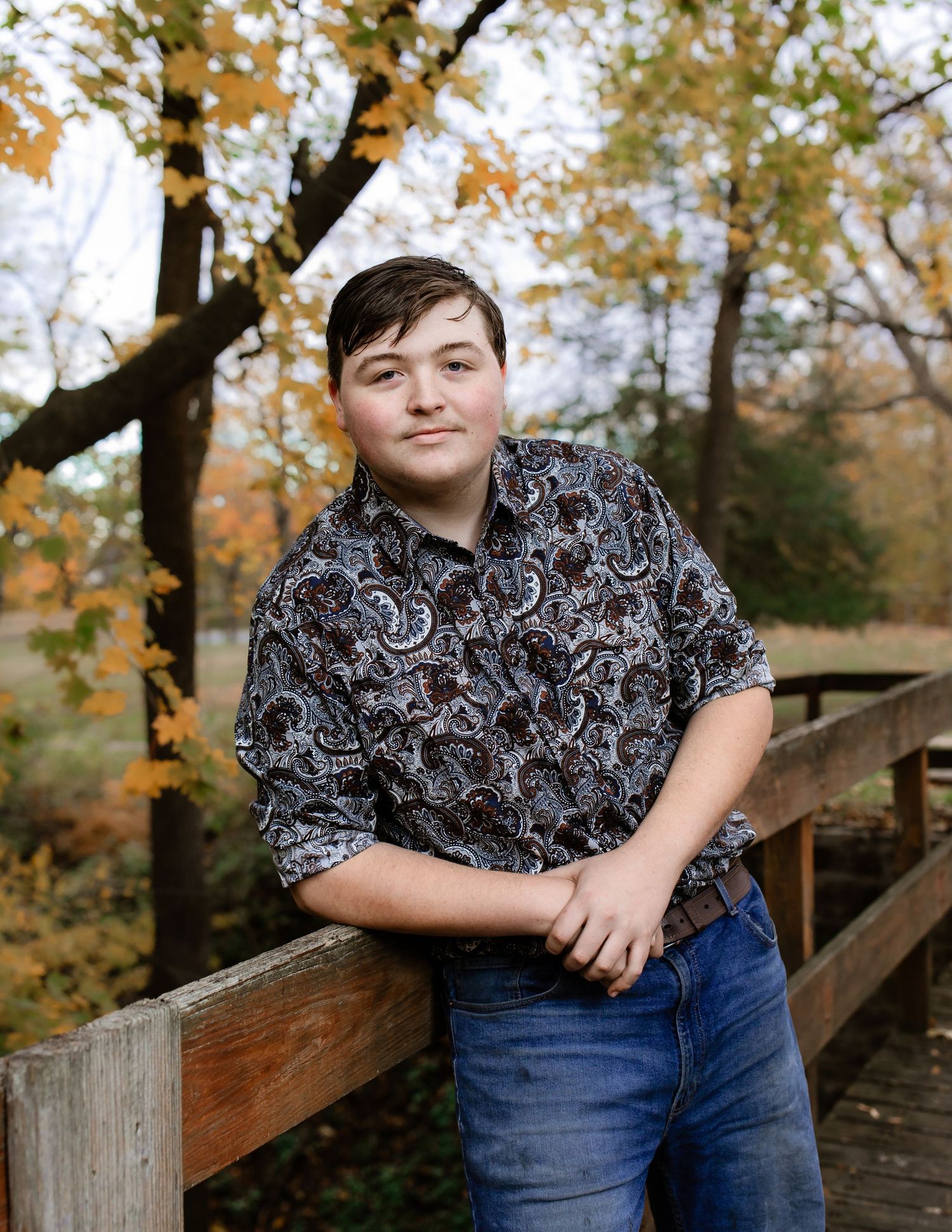 Young person in paisley print shirt and jeans poses near wooden railing during fall season.