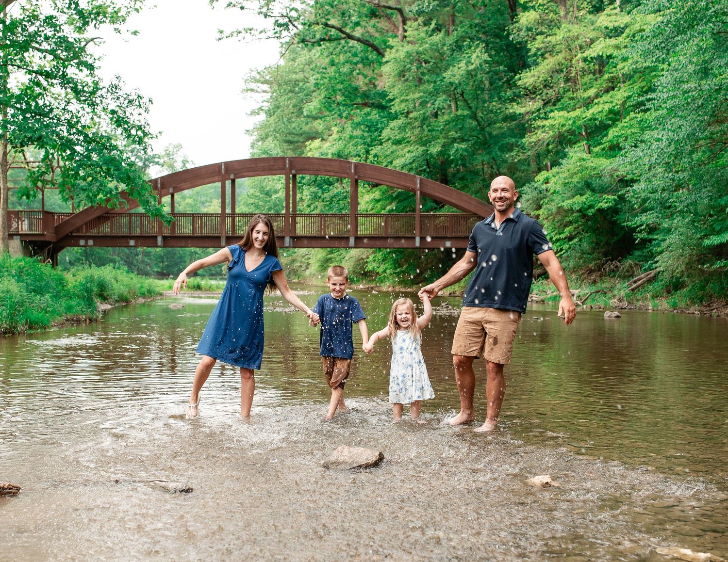 A family stands together in shallow creek water near a rustic red bridge surrounded by lush green trees.