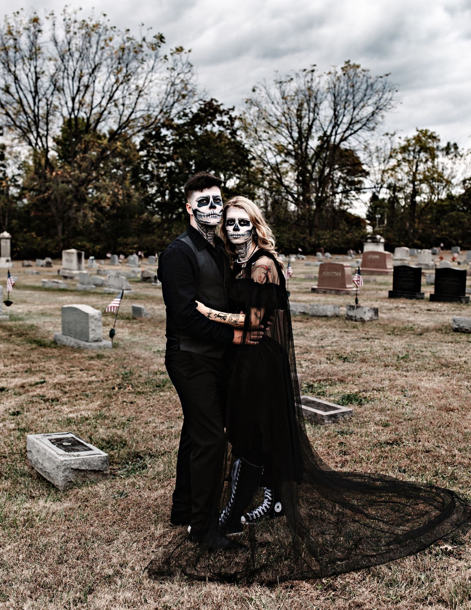 Gothic couple in black clothing poses dramatically in cemetery setting.