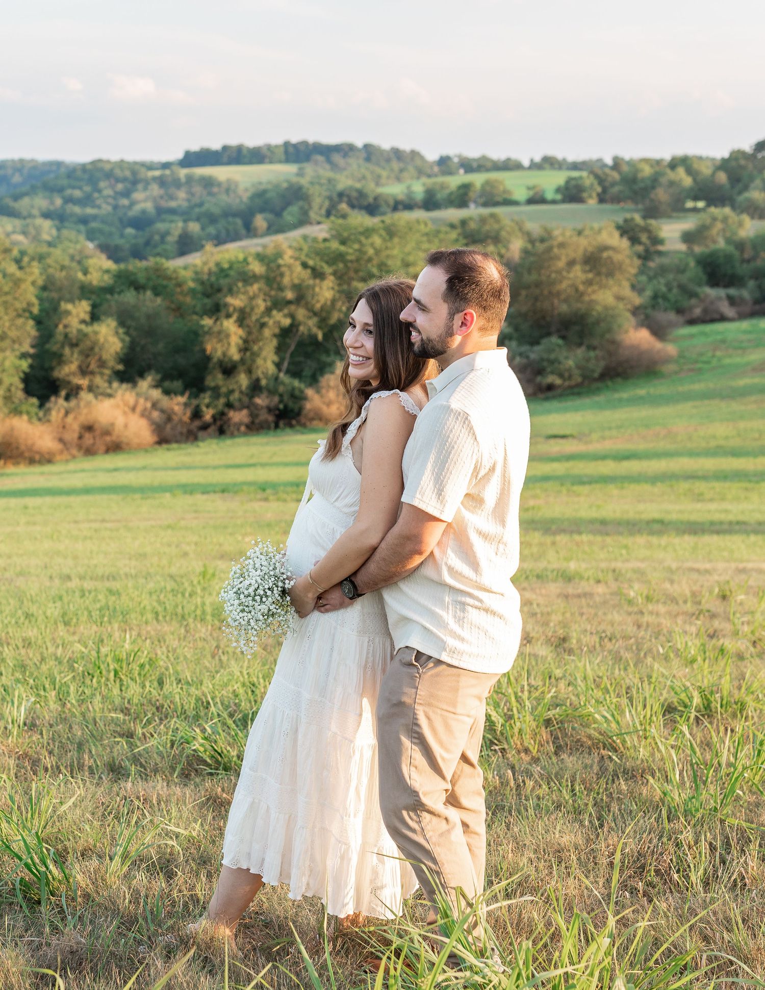 Pregnant couple poses in a sunny green field, woman in white dress holding baby's breath flowers, man embracing her from behind.