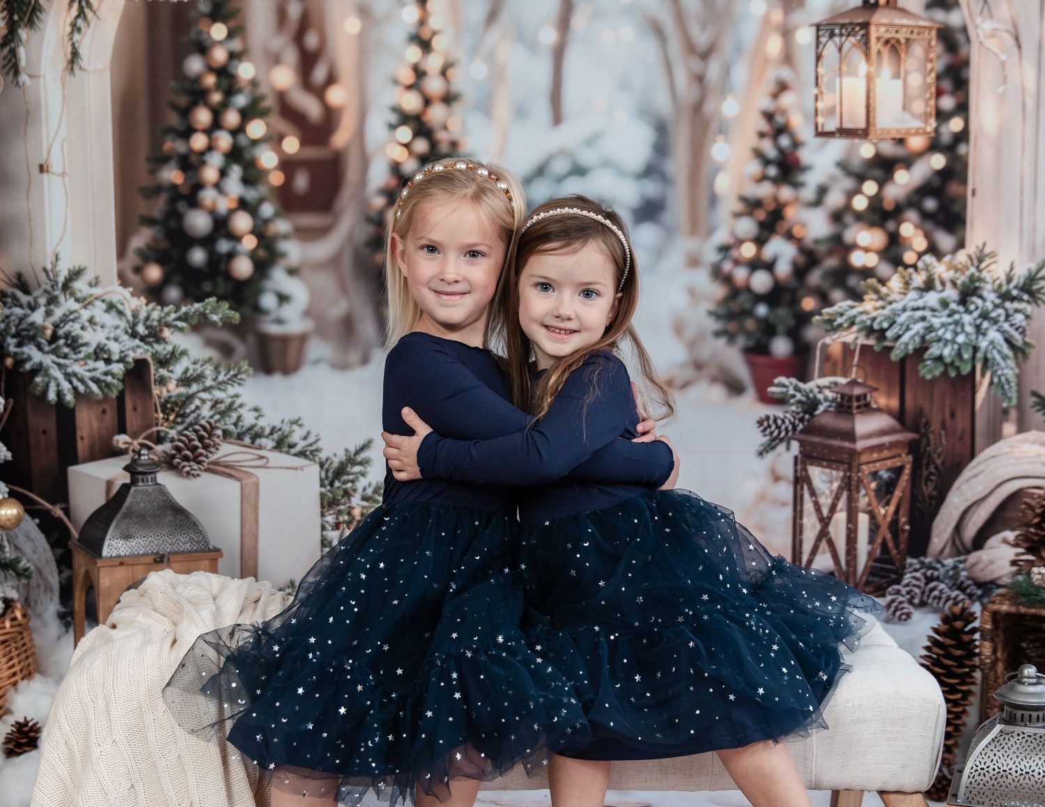 Two children in matching navy sparkly tulle dresses pose together in a festive holiday setting with Christmas trees and lanterns. Christmas