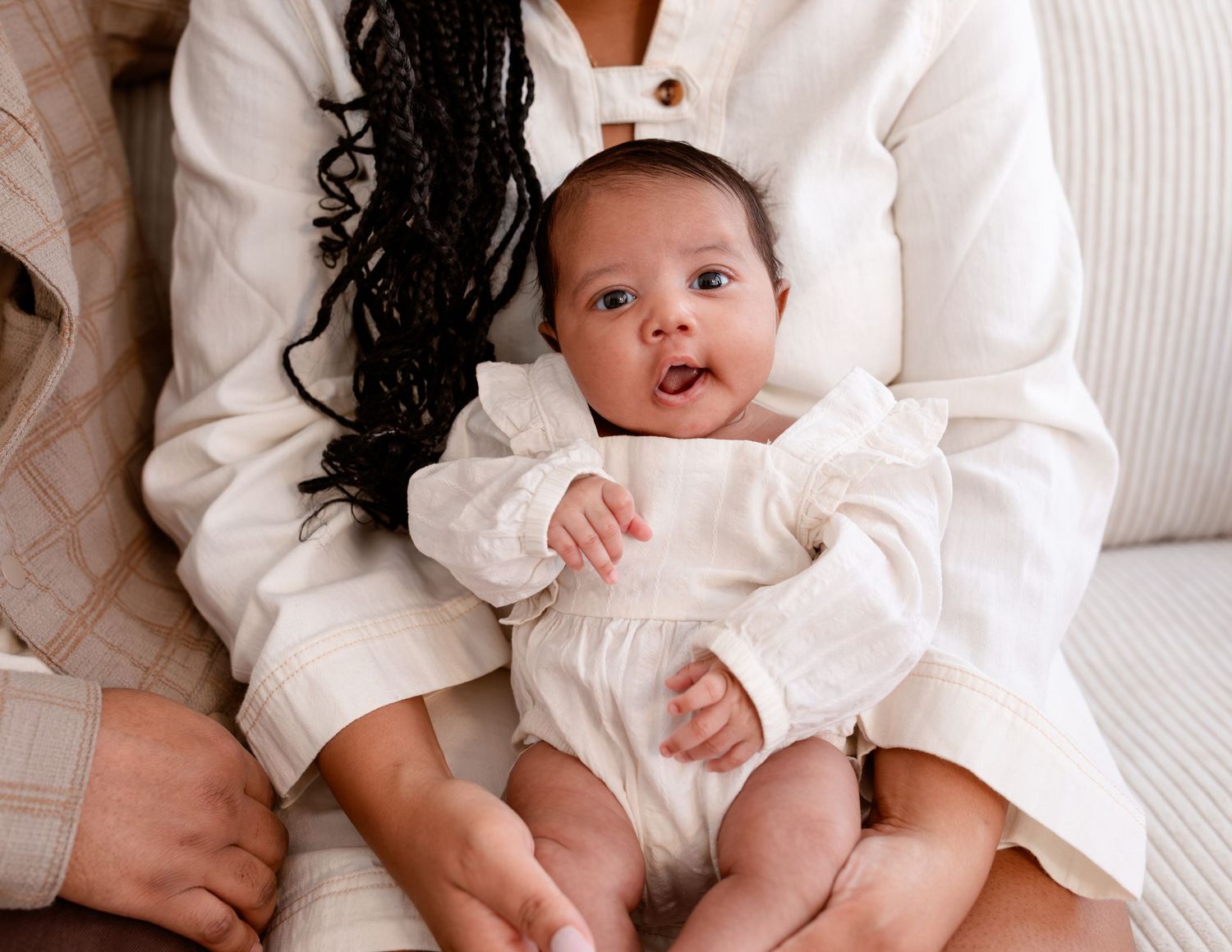 Baby wearing white outfit sits on lap with arms outstretched showing excitement and joy.