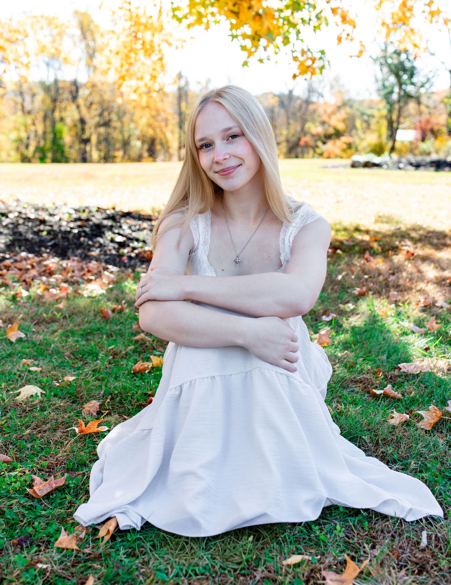 Young woman in a white dress sits on autumn grass surrounded by fallen leaves.