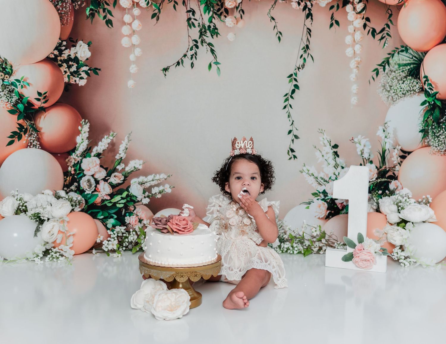 Floral cake smash setup with peach balloons, white flowers, and greenery against a soft pink backdrop.