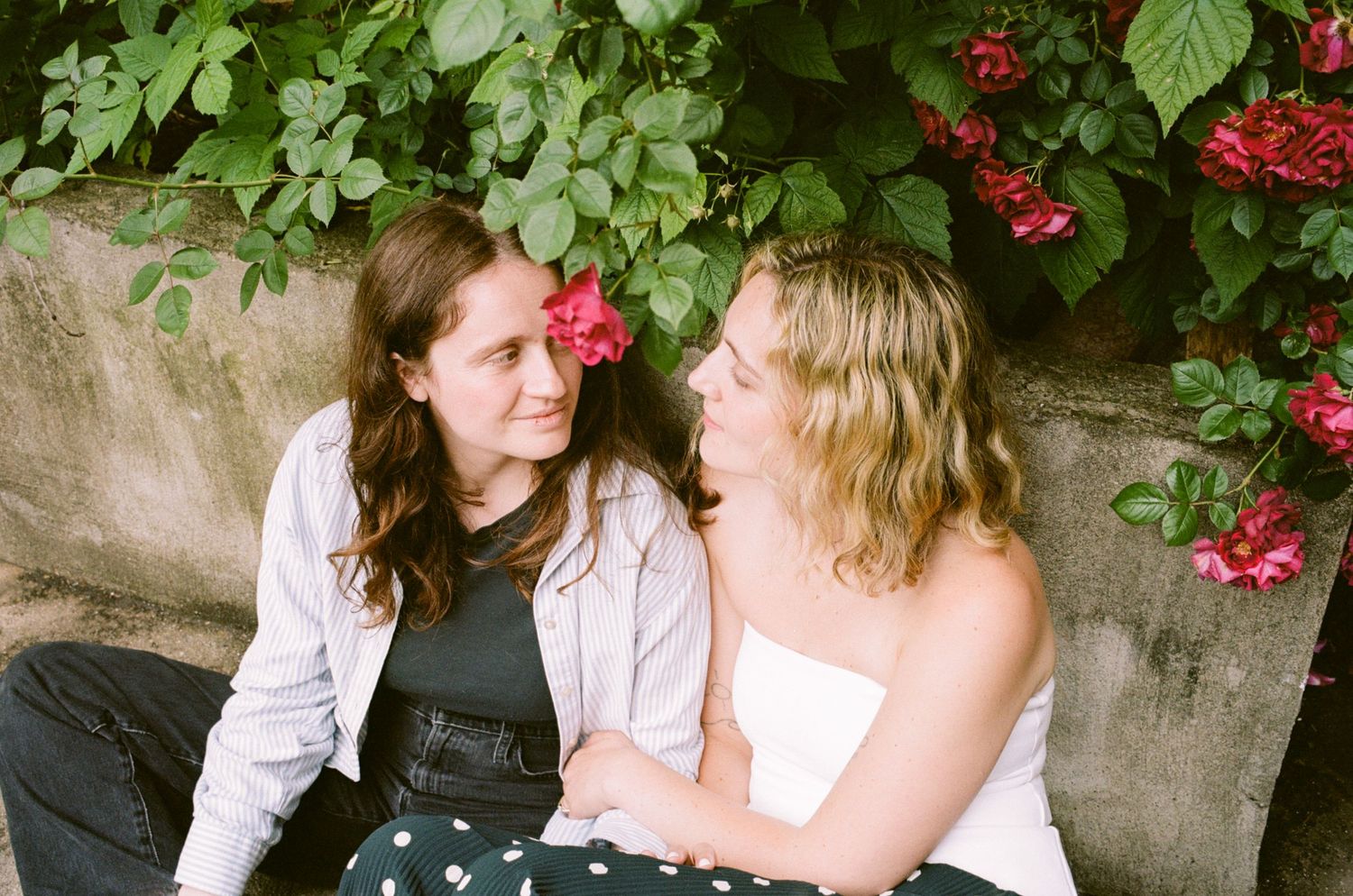 Two young women sitting together outdoors, smiling at each other, surrounded by lush green foliage and red roses.