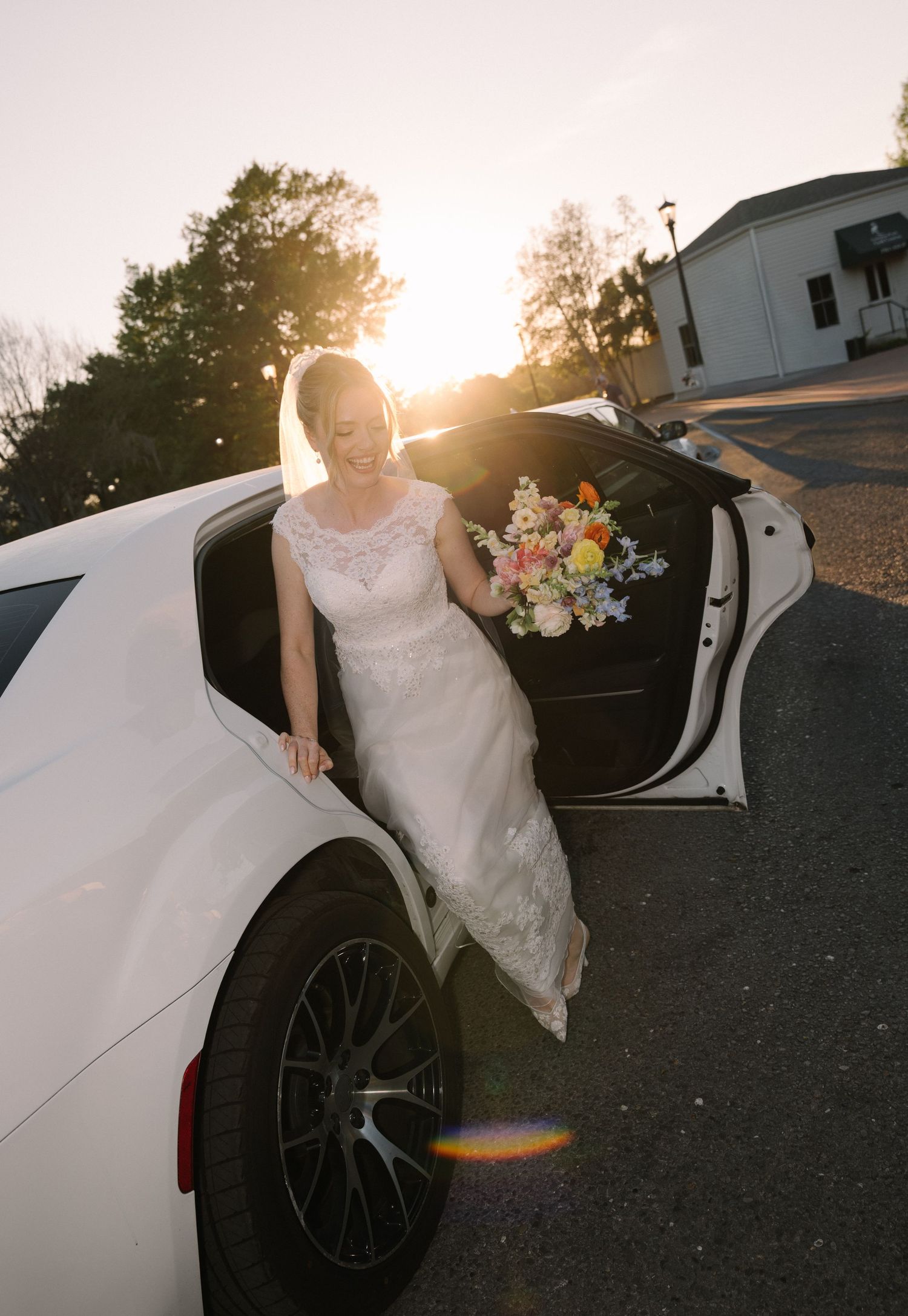 Bride in white lace gown holding colorful bouquet stepping out of white car at golden sunset.