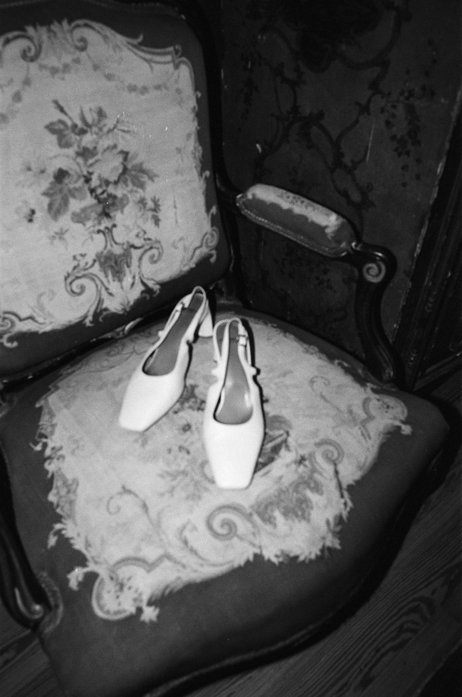 Black and white photo of white bridal shoes placed on an antique chair with floral upholstery.