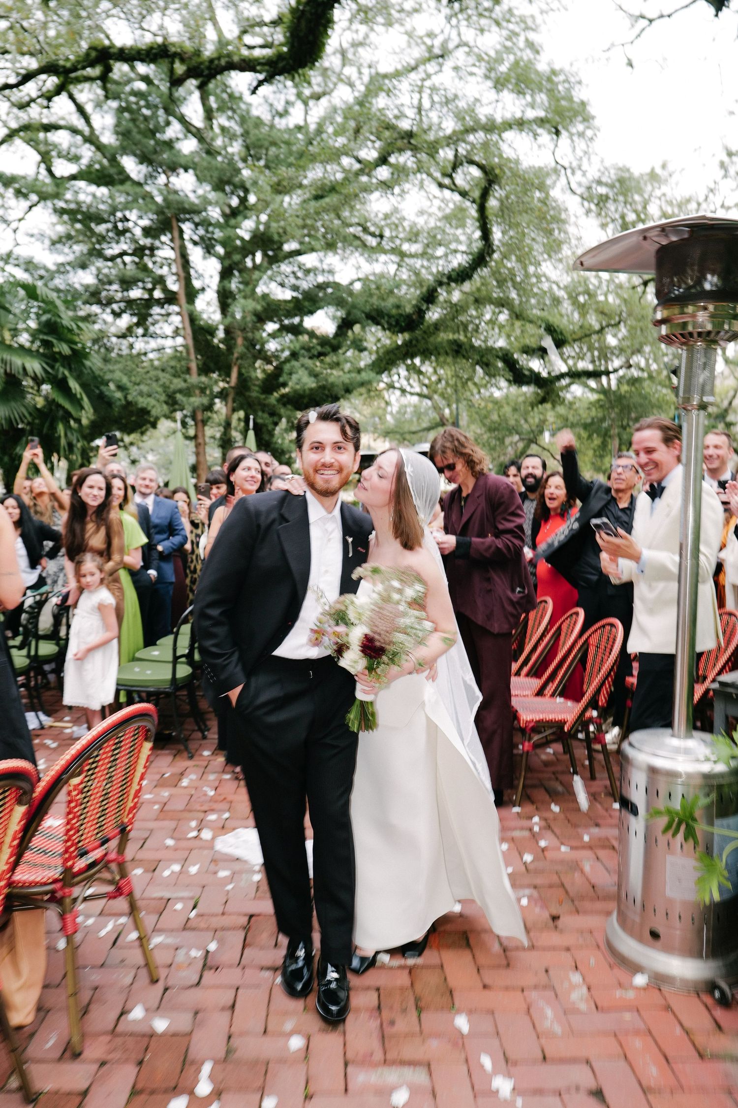 Wedding celebration on a brick patio surrounded by lush green trees.