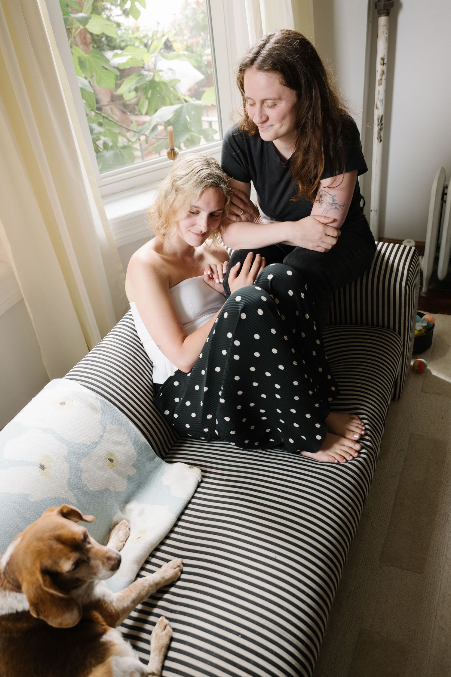 Two women relaxing on a striped chaise lounge with a beagle dog, near a bright window in a cozy home setting.