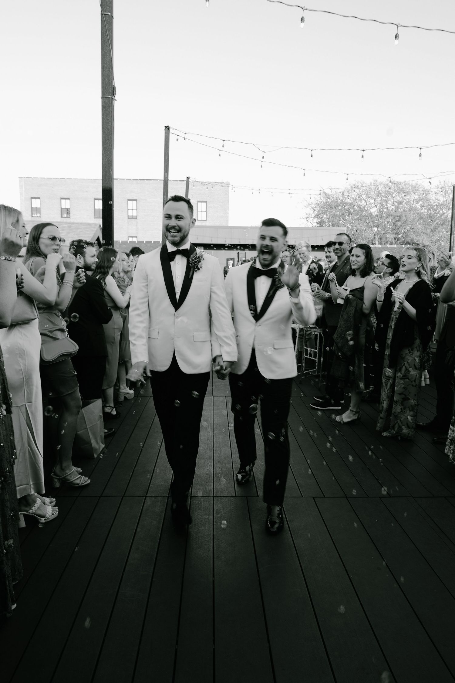 Wedding party in formal attire dancing in a line on outdoor deck with string lights overhead.