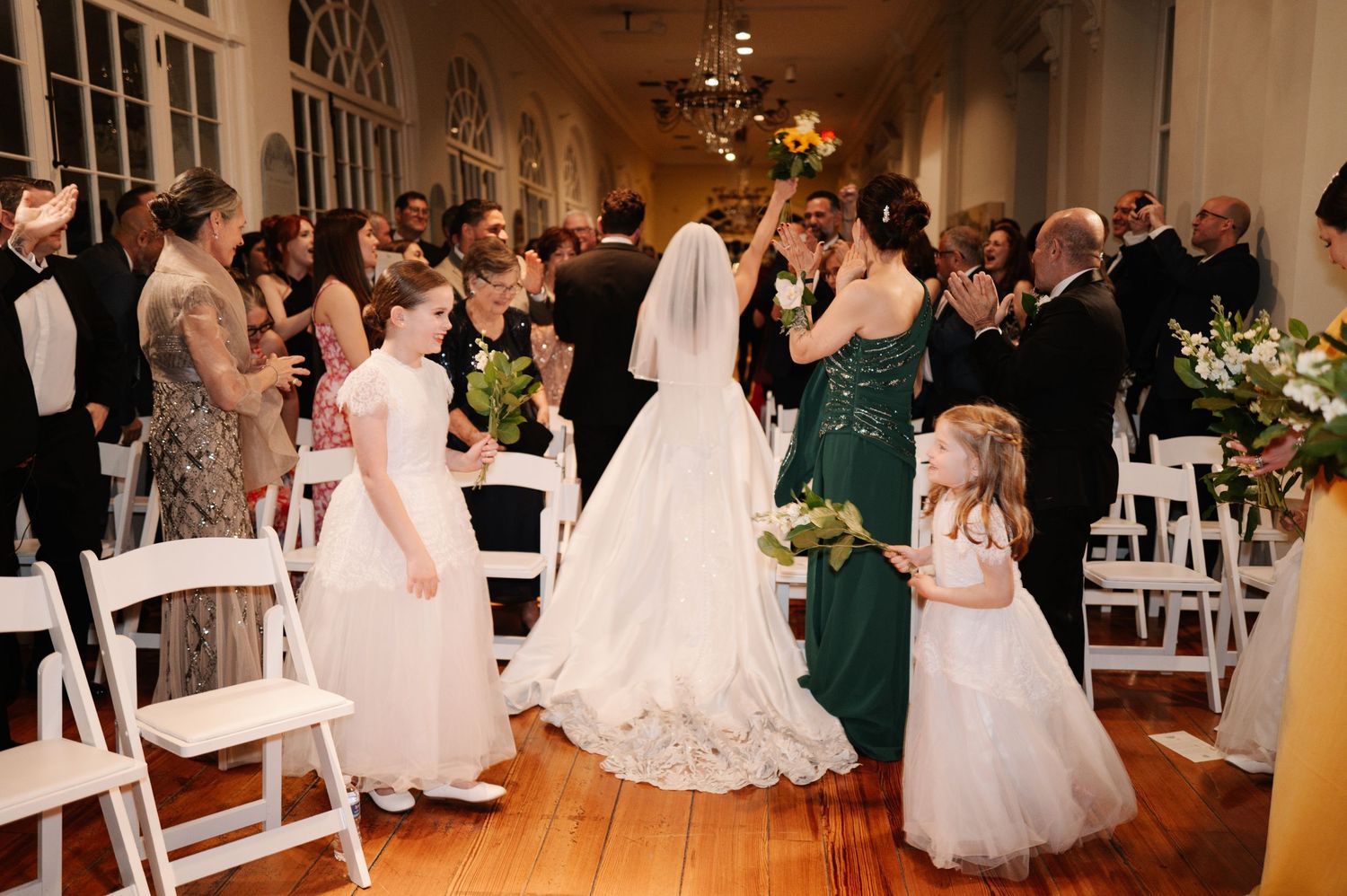 Bride in white gown walks down aisle with flower girls in white dresses at elegant indoor wedding ceremony.