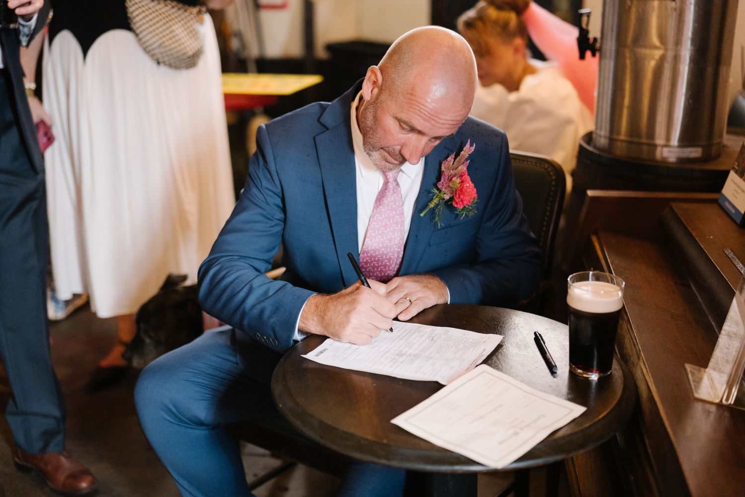 Groom in blue suit signing wedding documents at wooden table with glass of beer nearby.