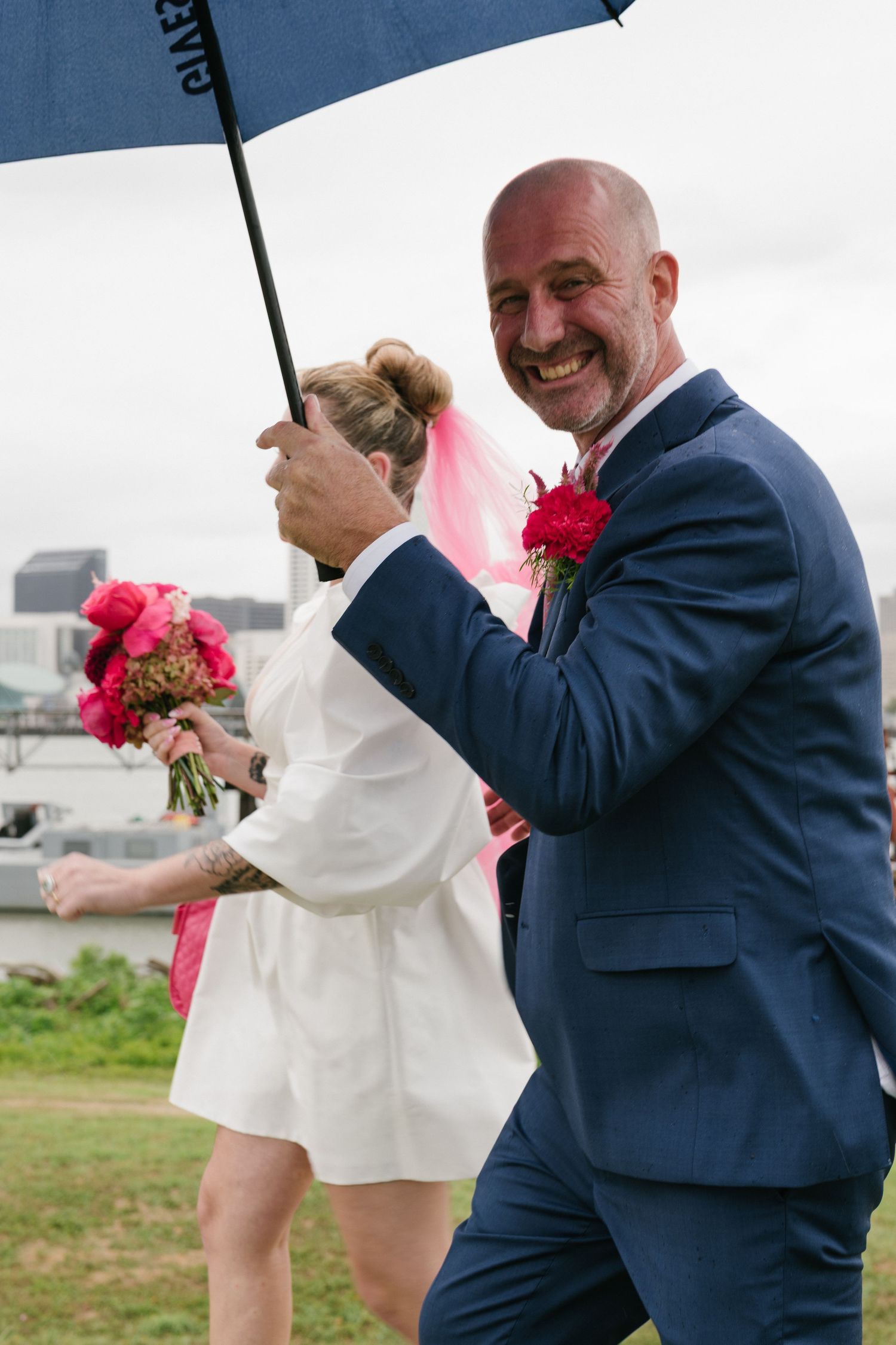 Groom in navy suit holds umbrella while walking outdoors with pink flowers.