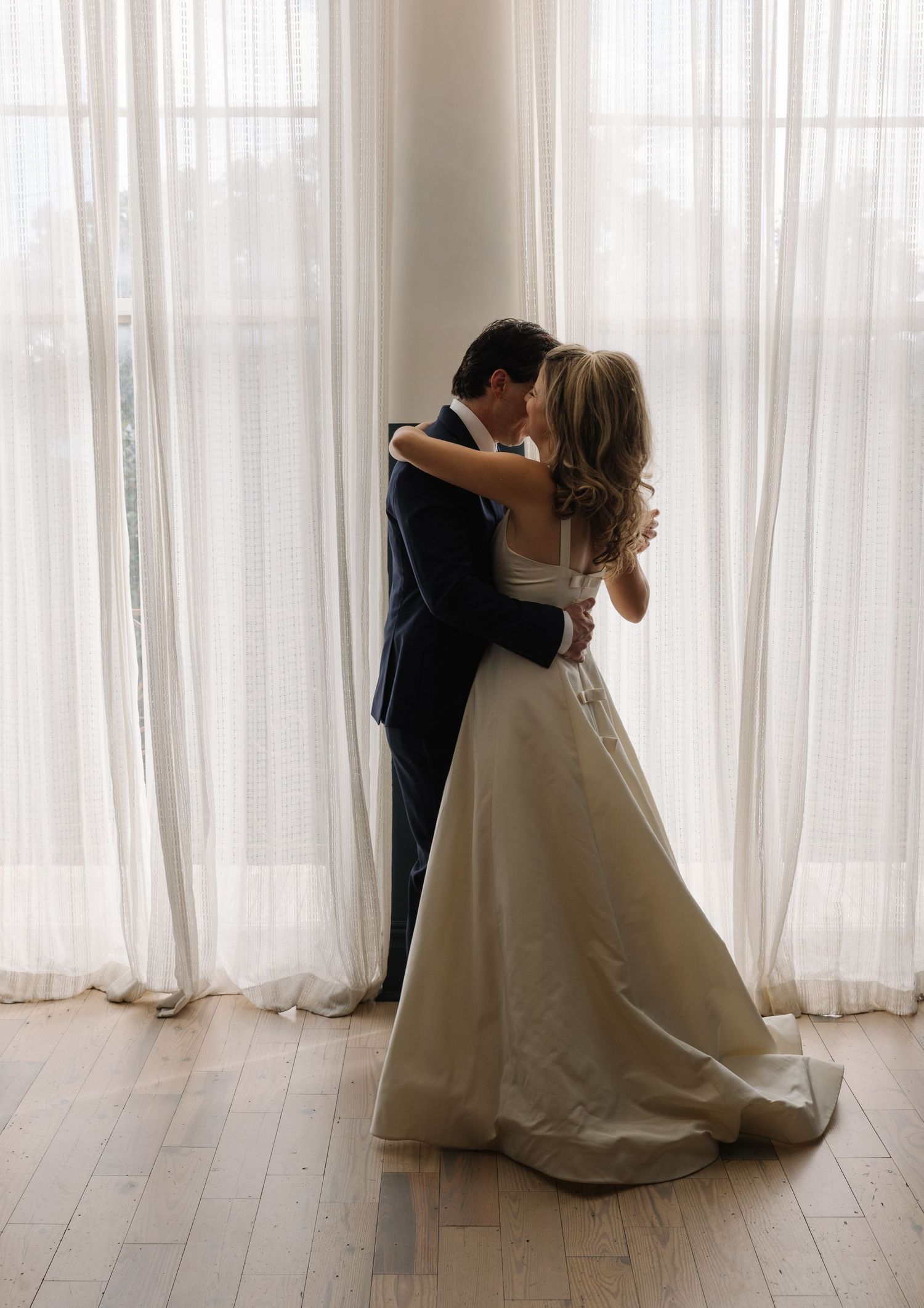 A bride and groom share a romantic kiss in front of sheer white curtains on their wedding day.