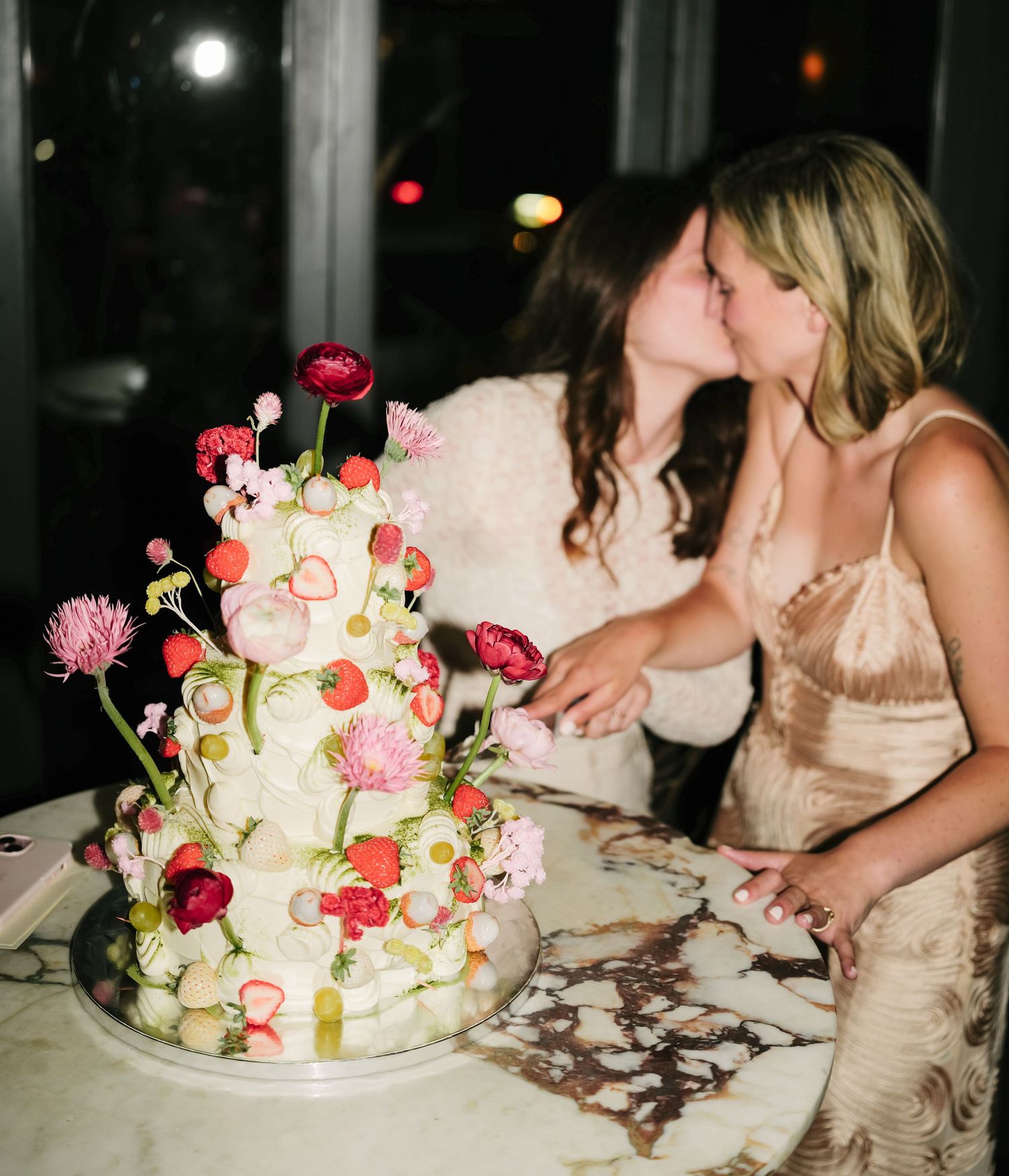 A decorative white wedding cake adorned with pink and red flowers sits in front of a married couple sharing a kiss.