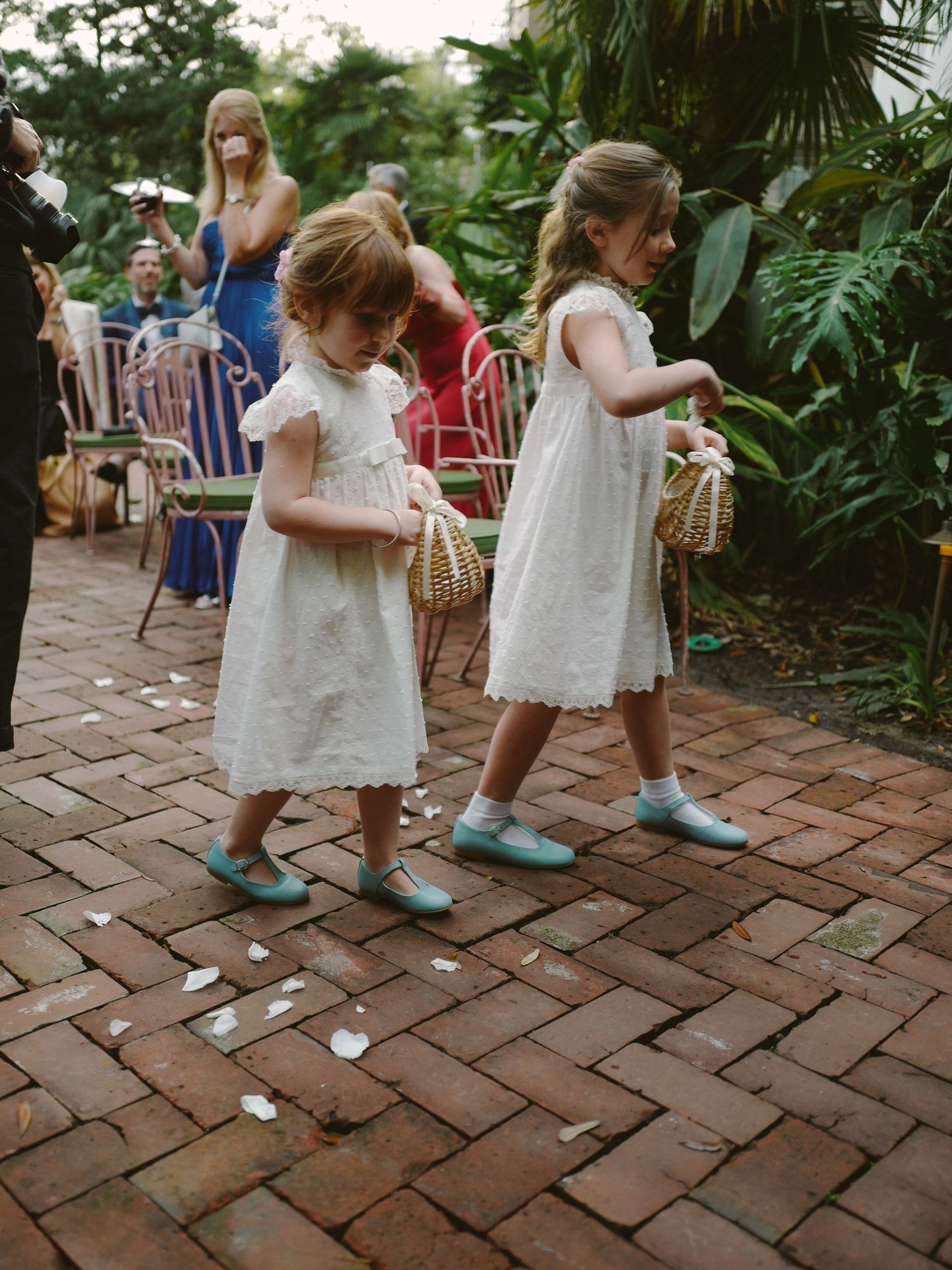 Flower girls in white dresses scattering flower petals along brick pathway.