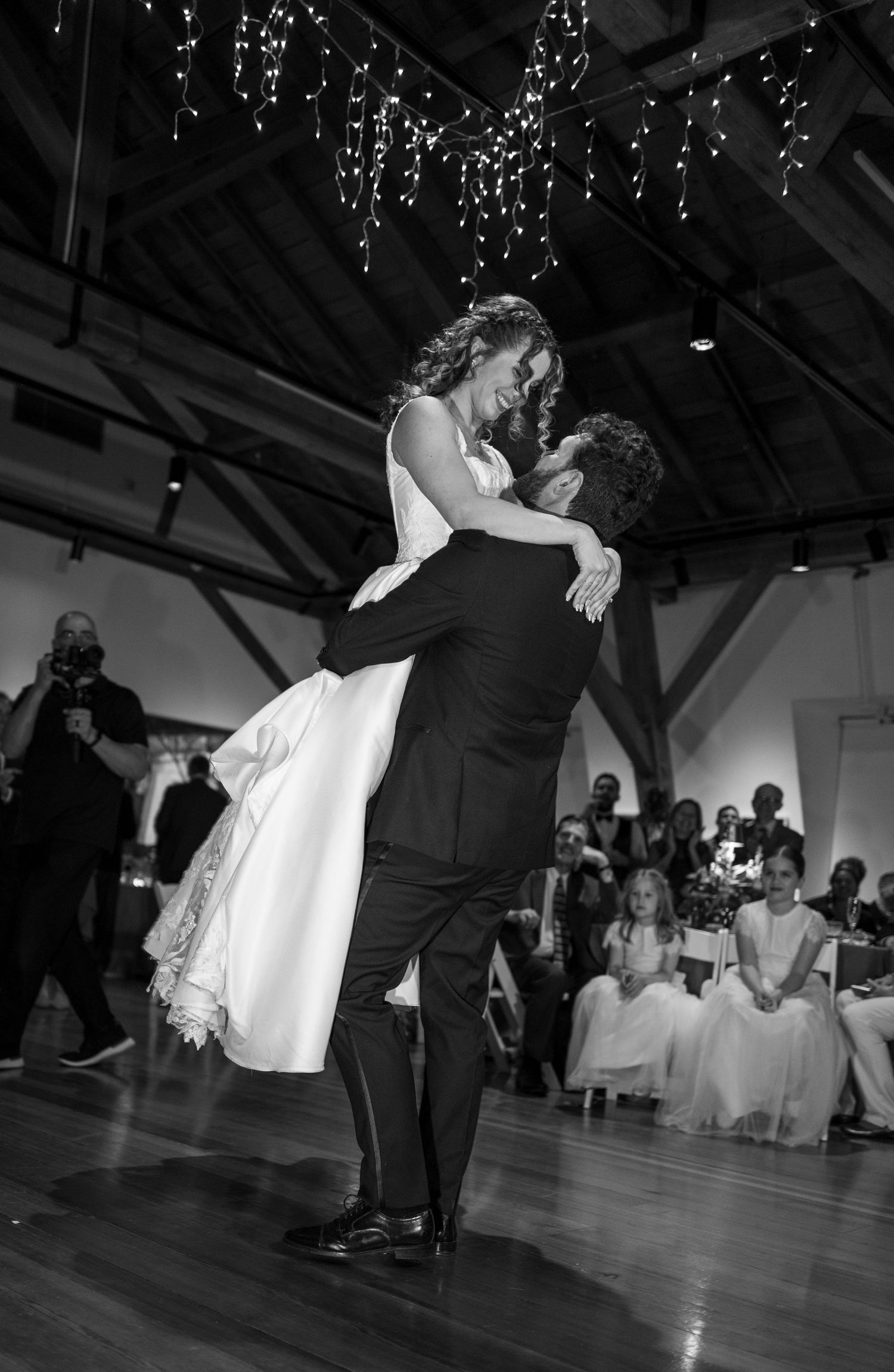 Groom lifts bride during first dance at wedding reception, black and white photo with fairy lights above.