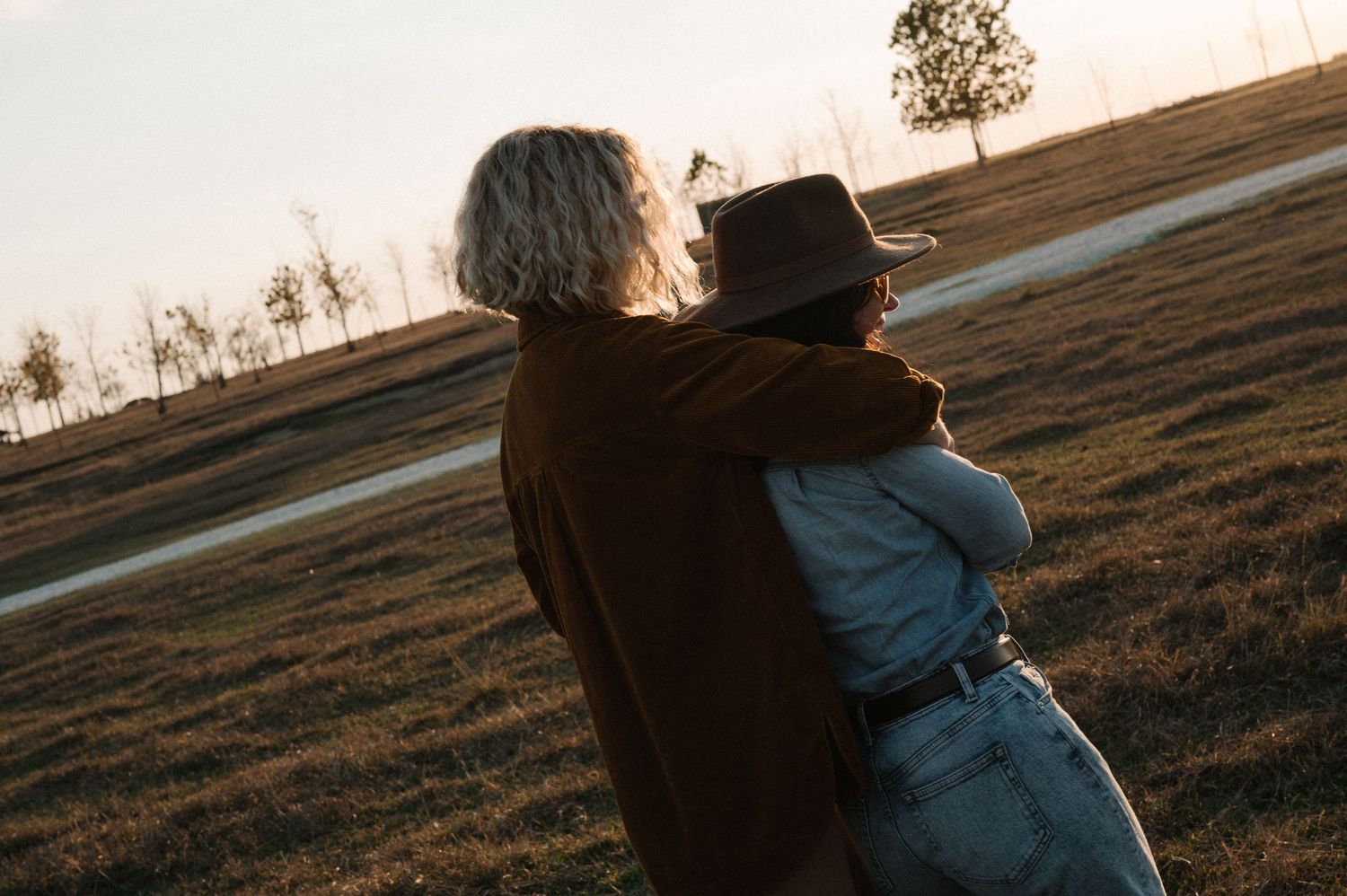 Two people embracing in an open field at sunset, one with white hair wearing a brown jacket.