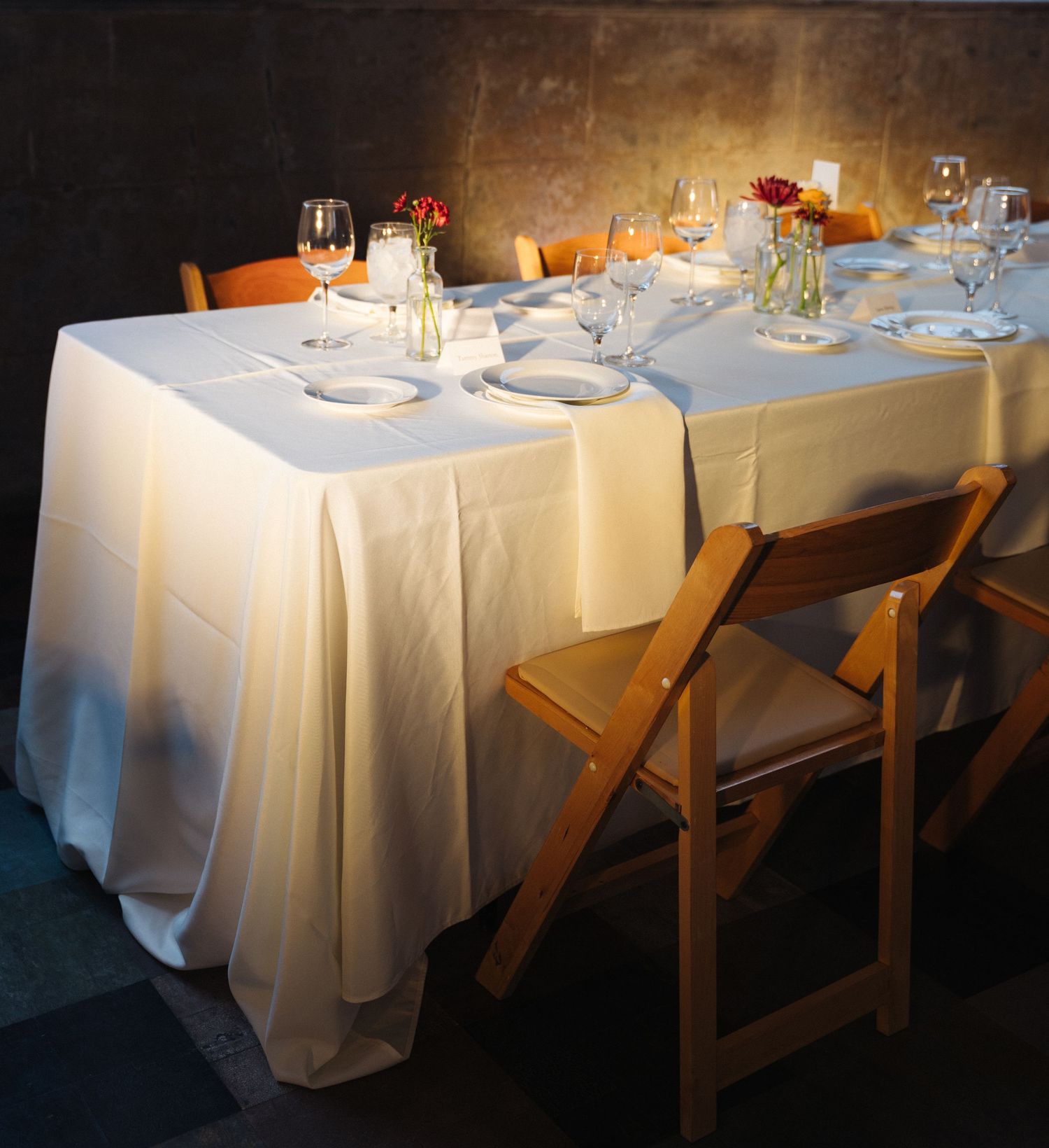 Elegant table setting with white linens, wine glasses and wooden chairs in warm lighting.