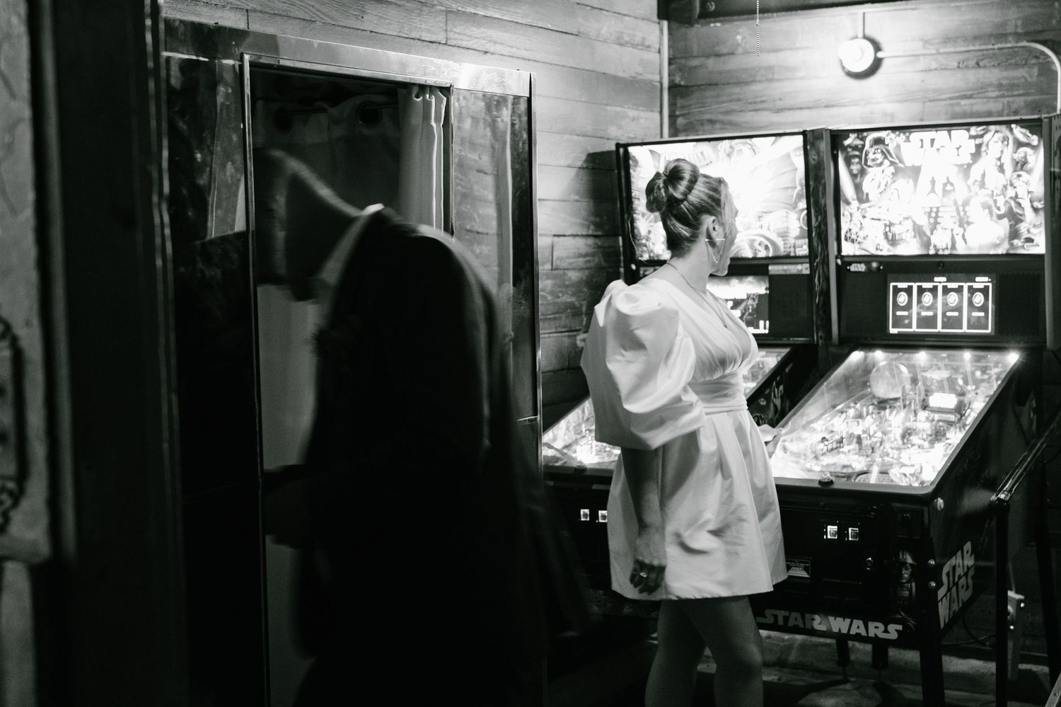 Black and white photo of a woman in a white dress playing a vintage pinball machine in a dimly lit arcade room.