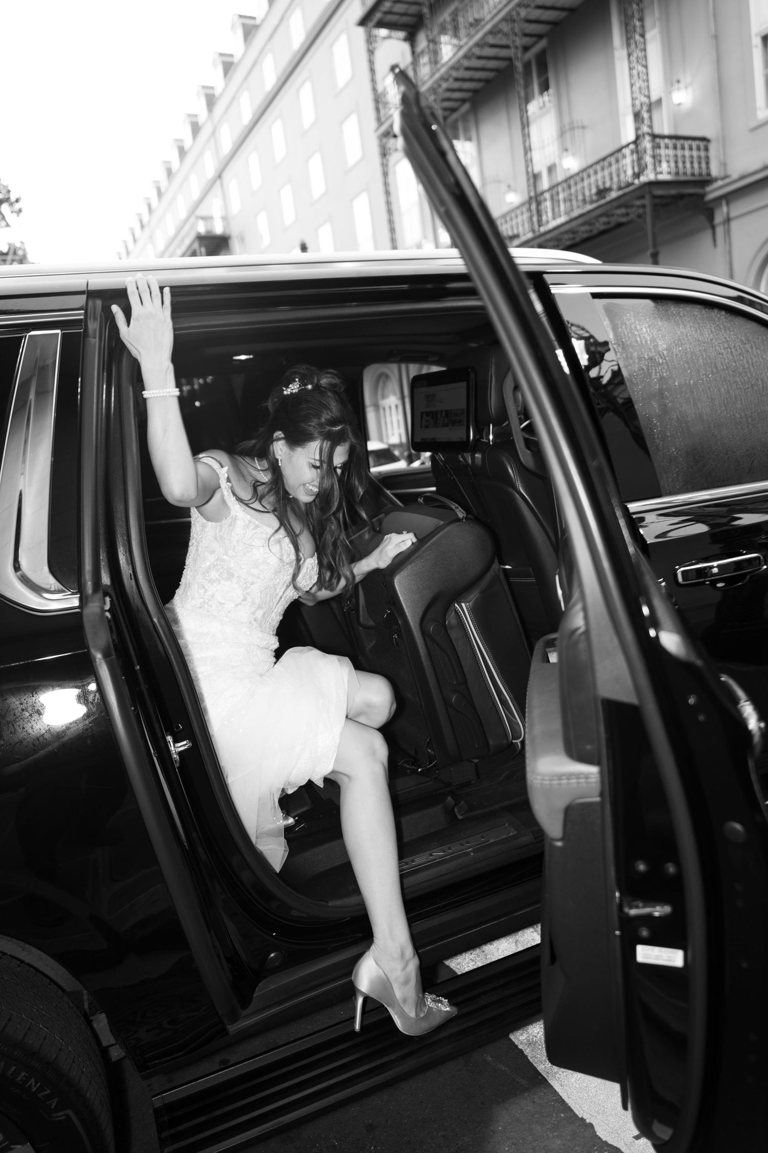 A bride in a white dress exits a vintage black car while wearing high heels.