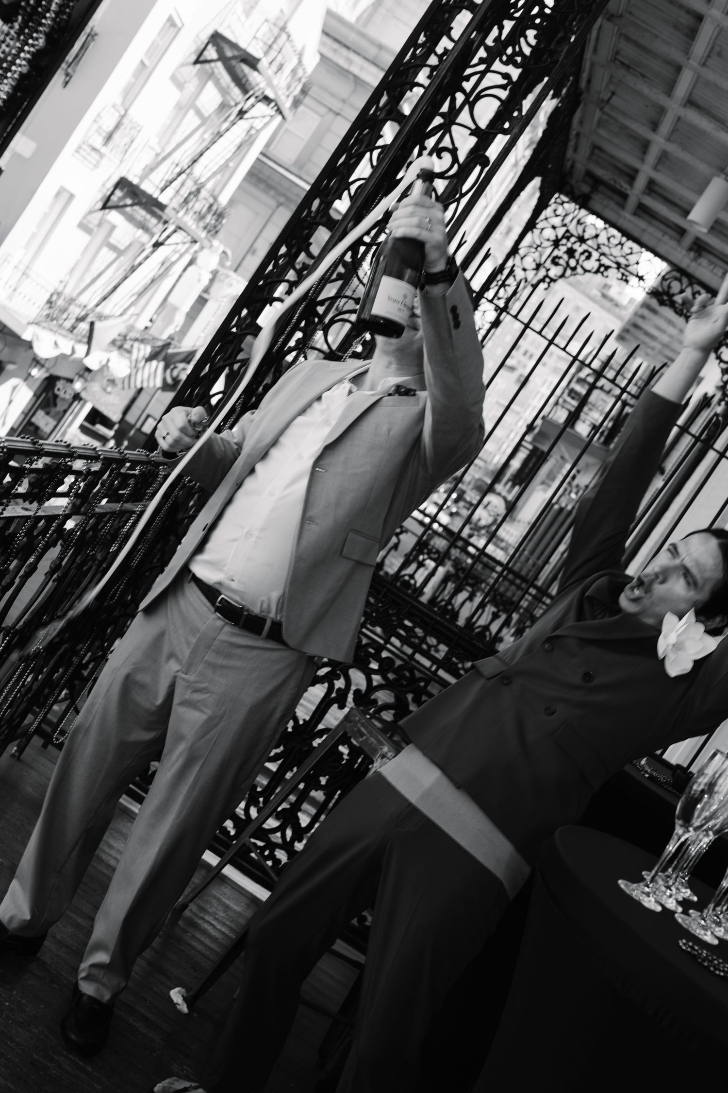 Black and white photo of a man in a suit raising a bottle at a social gathering near ornate iron railings.