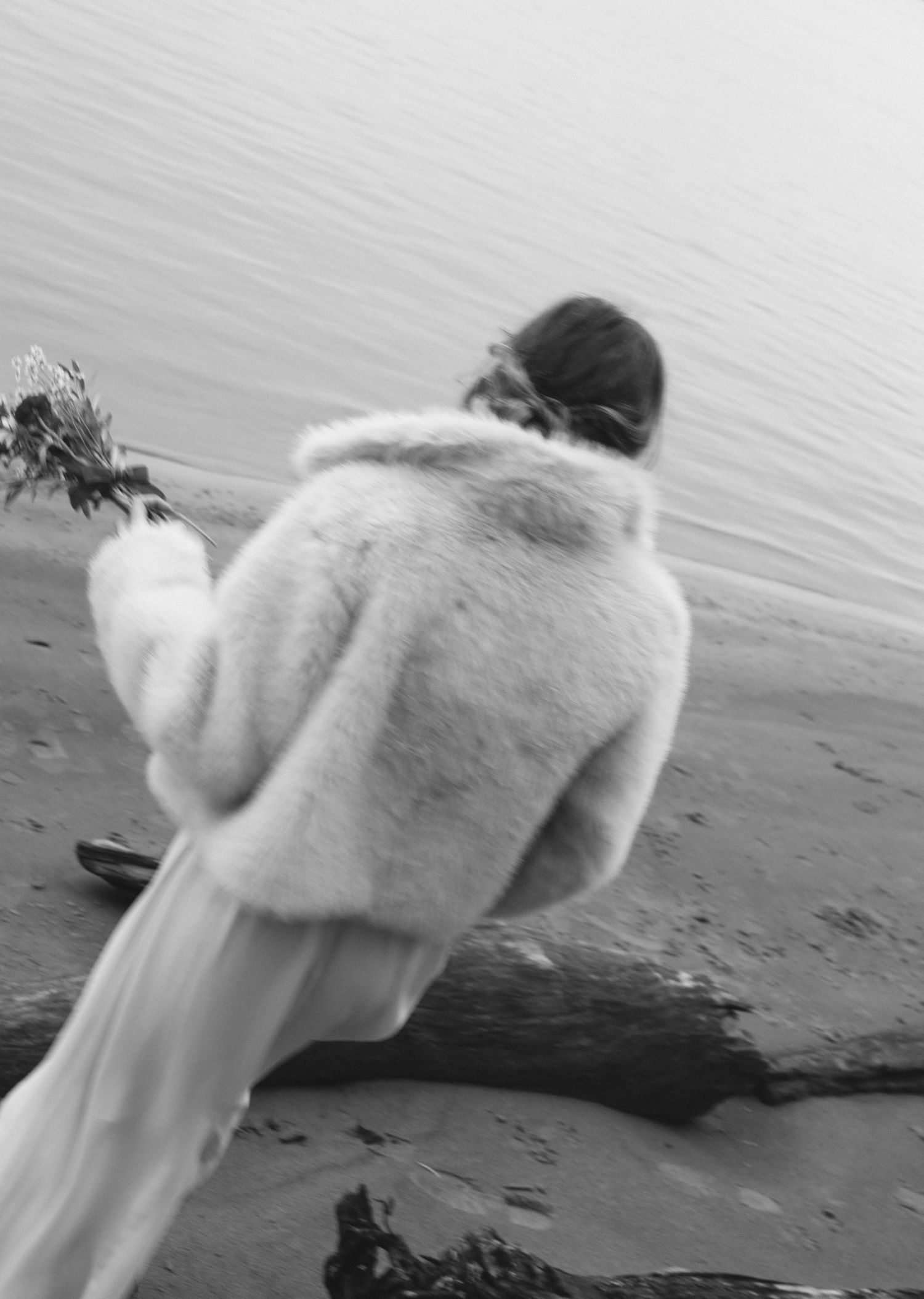 Black and white photo of a woman in a white fur coat standing on a rocky beach near driftwood and calm water.