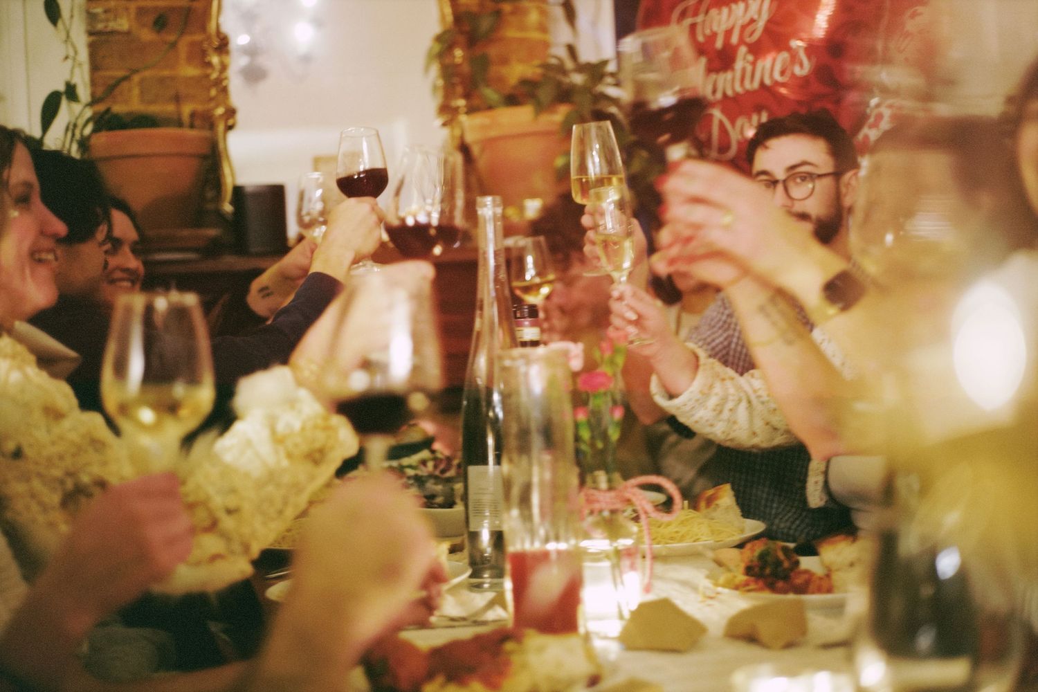 Friends raise wine glasses in a toast at a dimly lit rehearsal dinner party with warm vintage lighting.