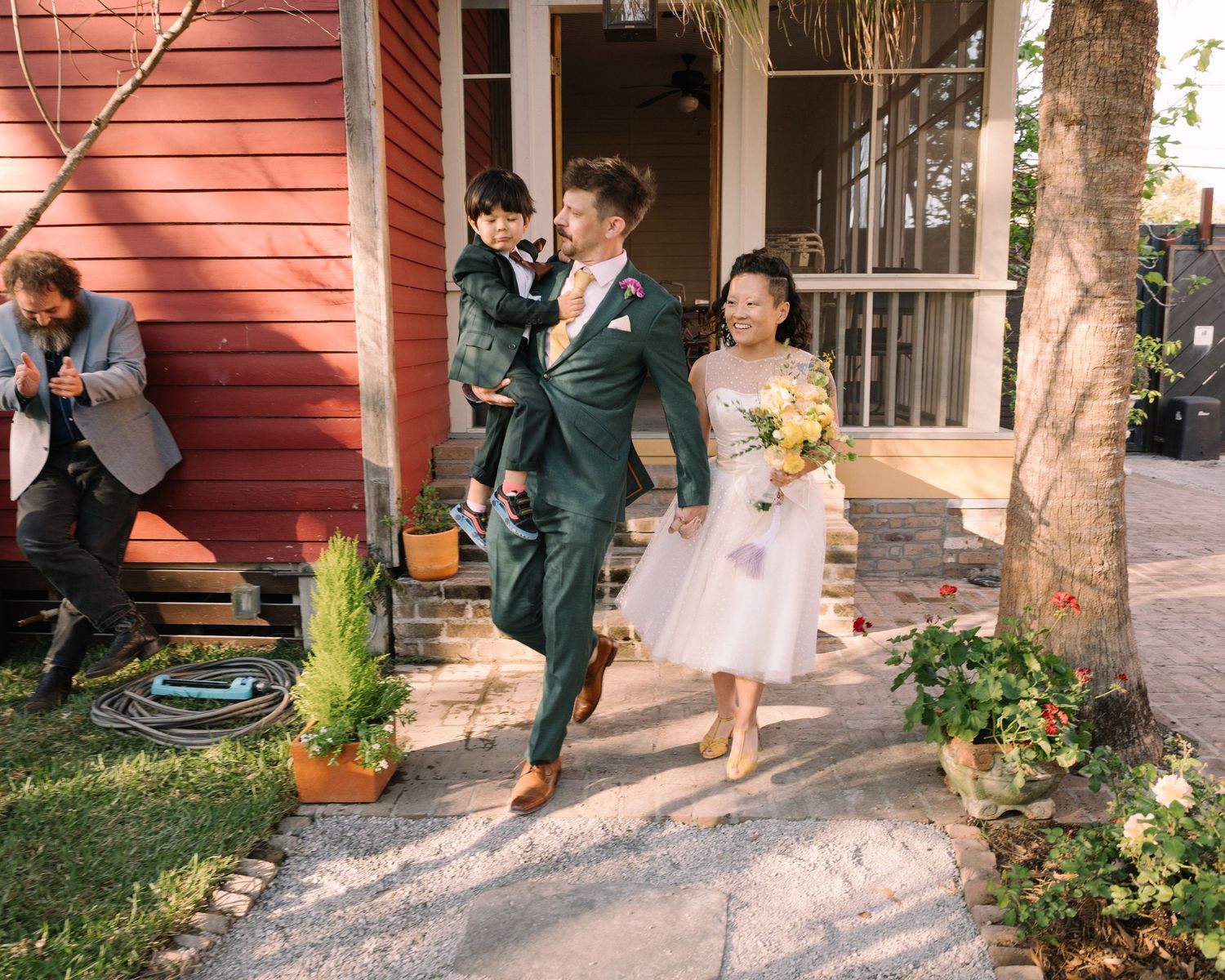 Joyful newlywed couple walking outside a red building, bride in white dress holding bouquet, groom in green suit.