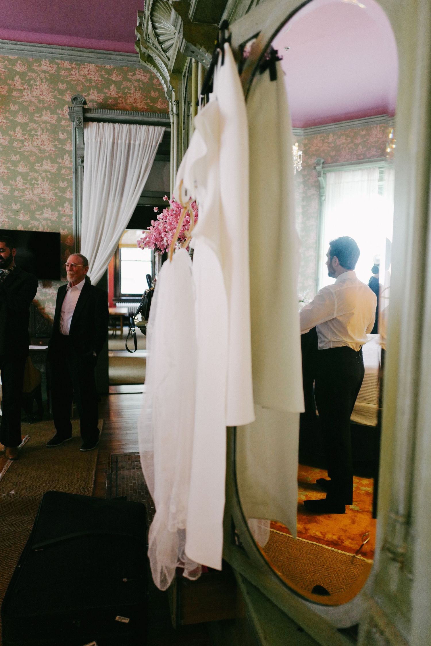 Wedding dress hanging in an elegant room with ornate mirror and floral wallpaper.