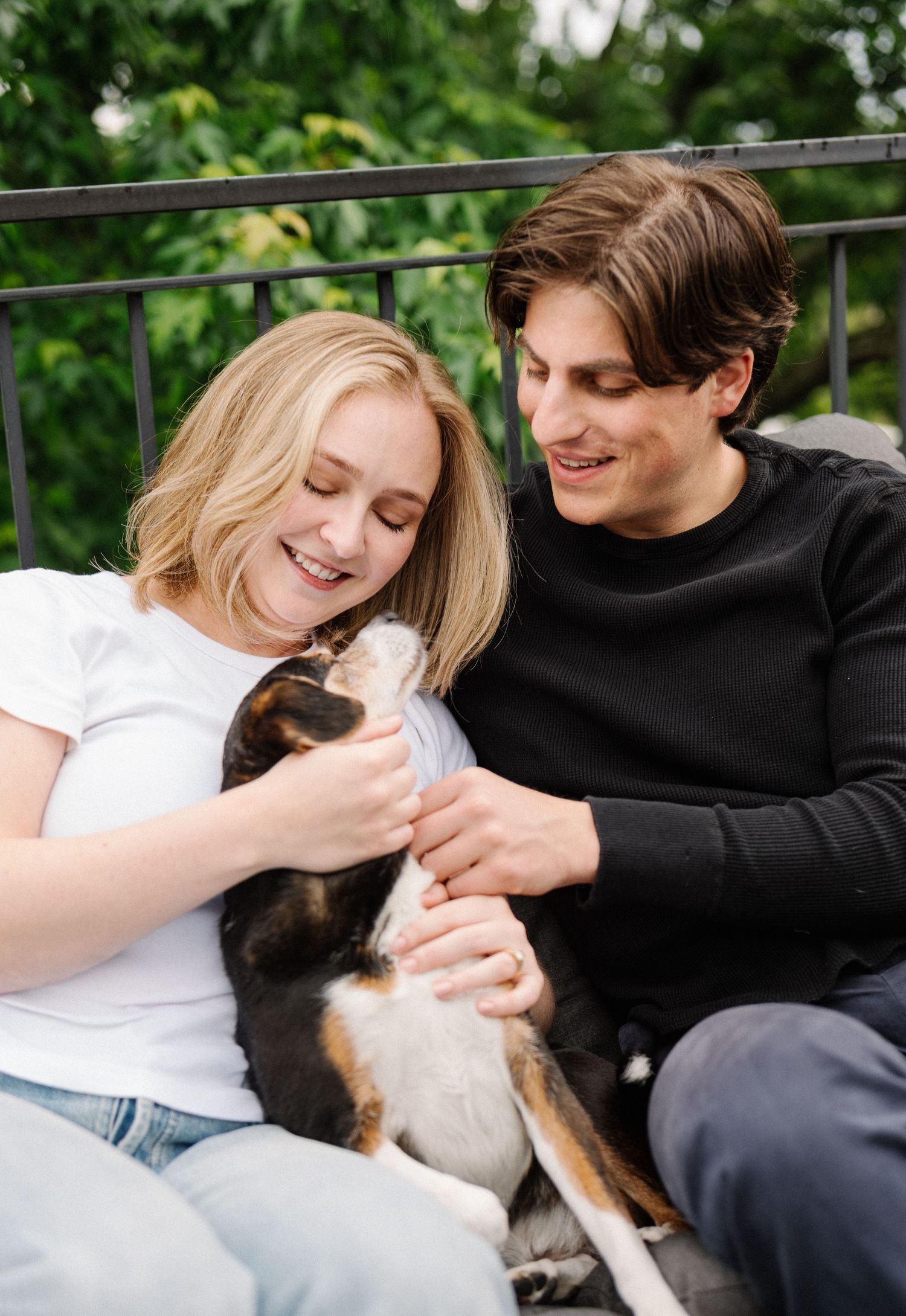 A smiling young couple sitting outdoors on a balcony, happily petting a small tricolor puppy together.