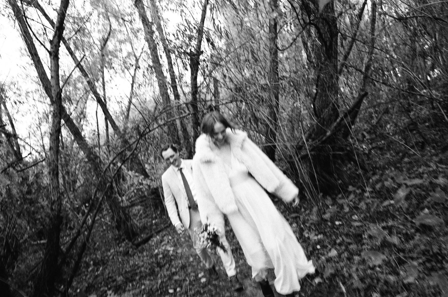 Black and white photo of a bride in white gown and groom in suit walking through a wooded forest area.