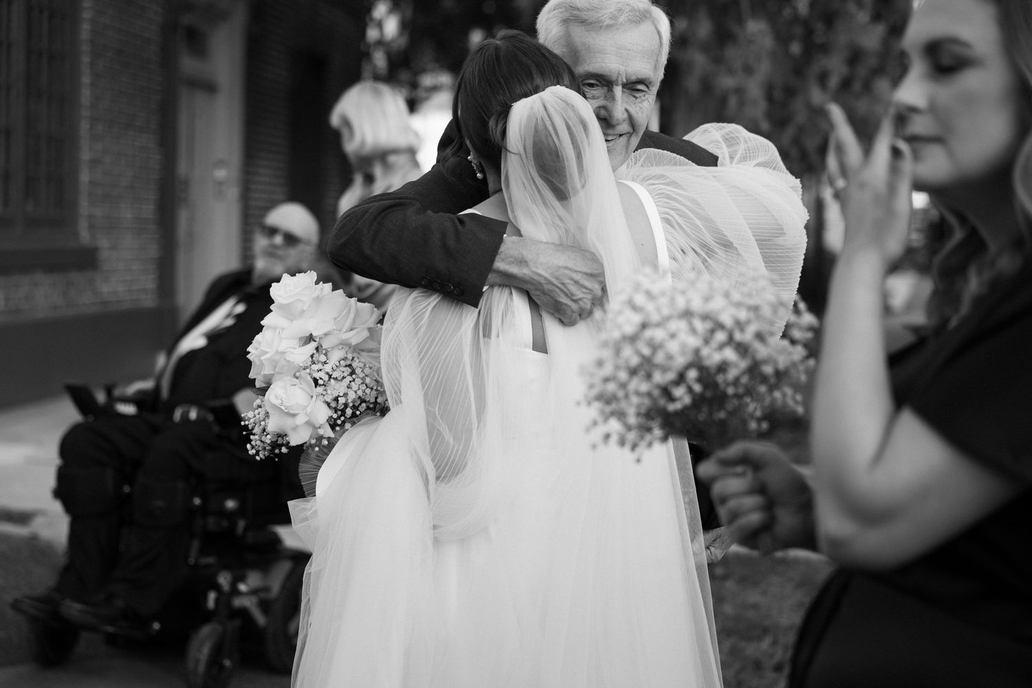 Black and white wedding photo of bride hugging older man, holding bouquet, with guests in background.