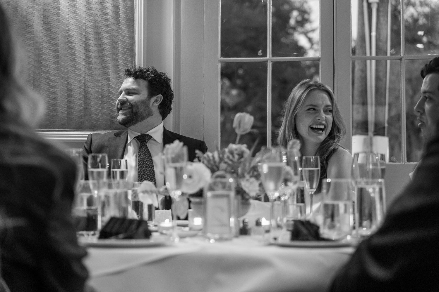Black and white photo of a laughing couple at a wedding reception dinner table with elegant glassware.