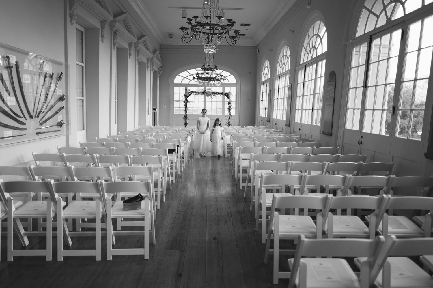 Black and white photo of an elegant wedding ceremony hall with white chairs, arched windows, and a chandelier.